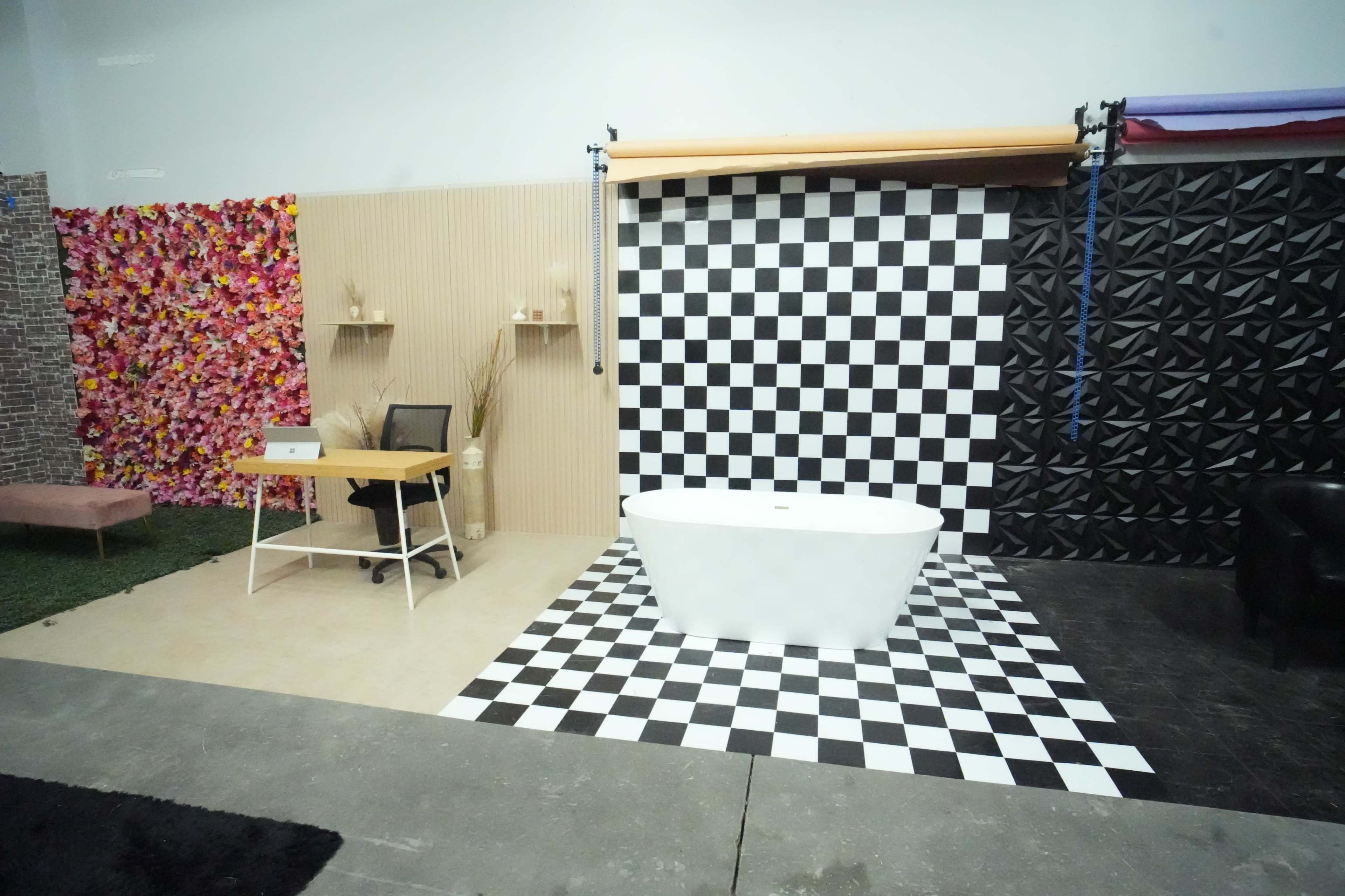 The image shows a studio space featuring a white freestanding bathtub on a black-and-white checkerboard floor, with decorative wall panels in the background and a desk set up in one corner.
