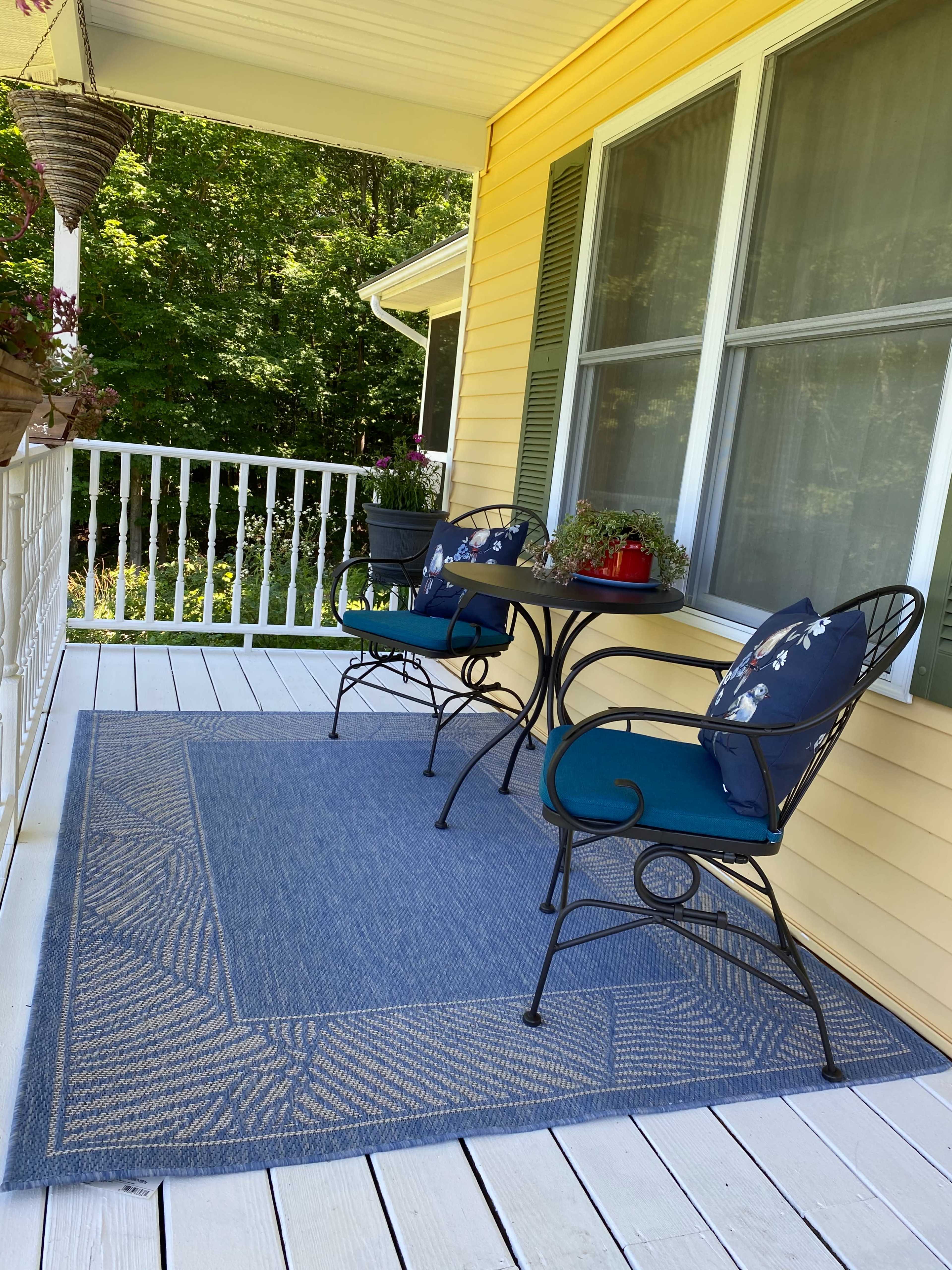 A small porch with two metal chairs and a round table, surrounded by a blue rug, with potted plants and a view of greenery in the background.
