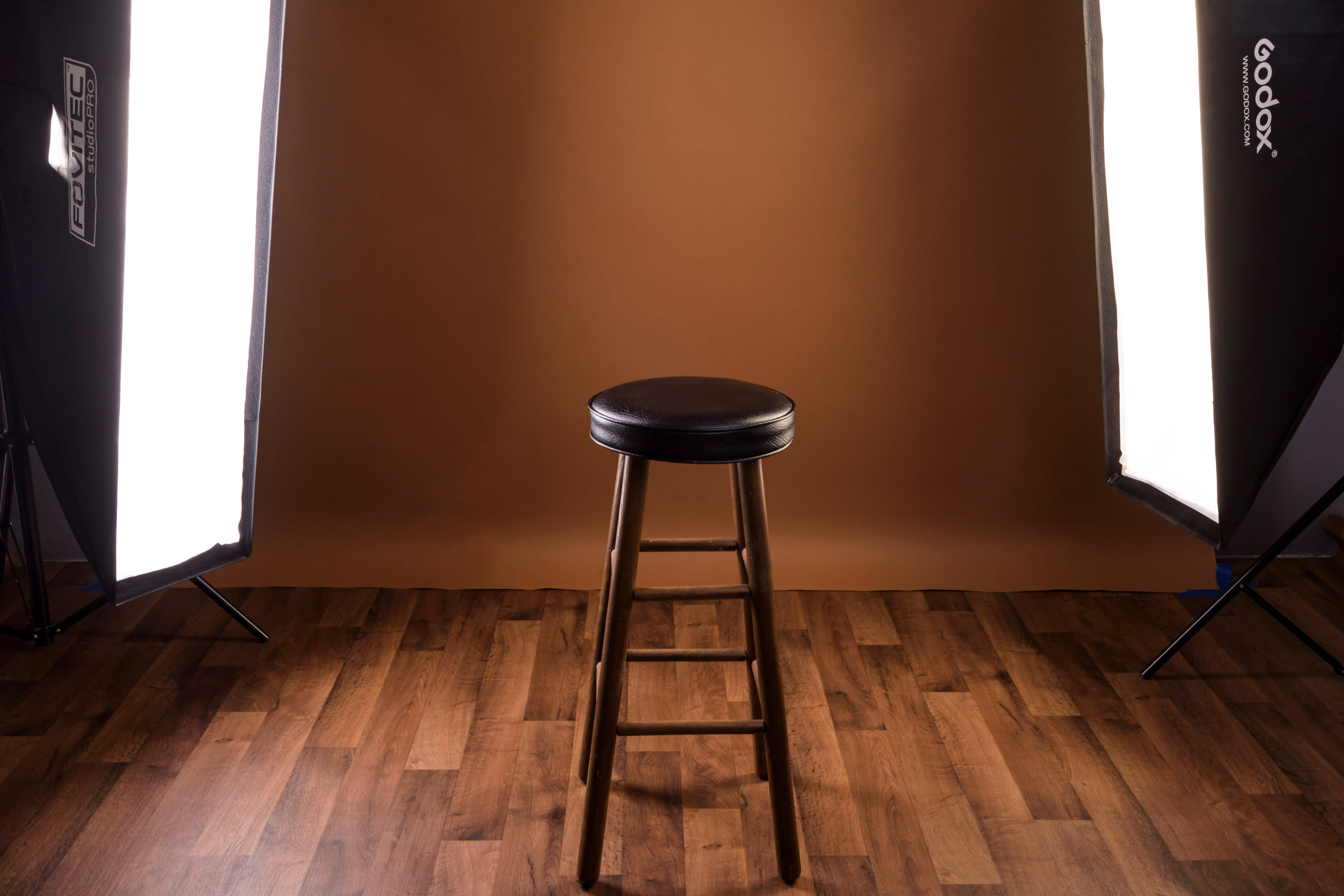 A black stool stands in the center of a well-lit studio with a brown backdrop and wooden flooring.
