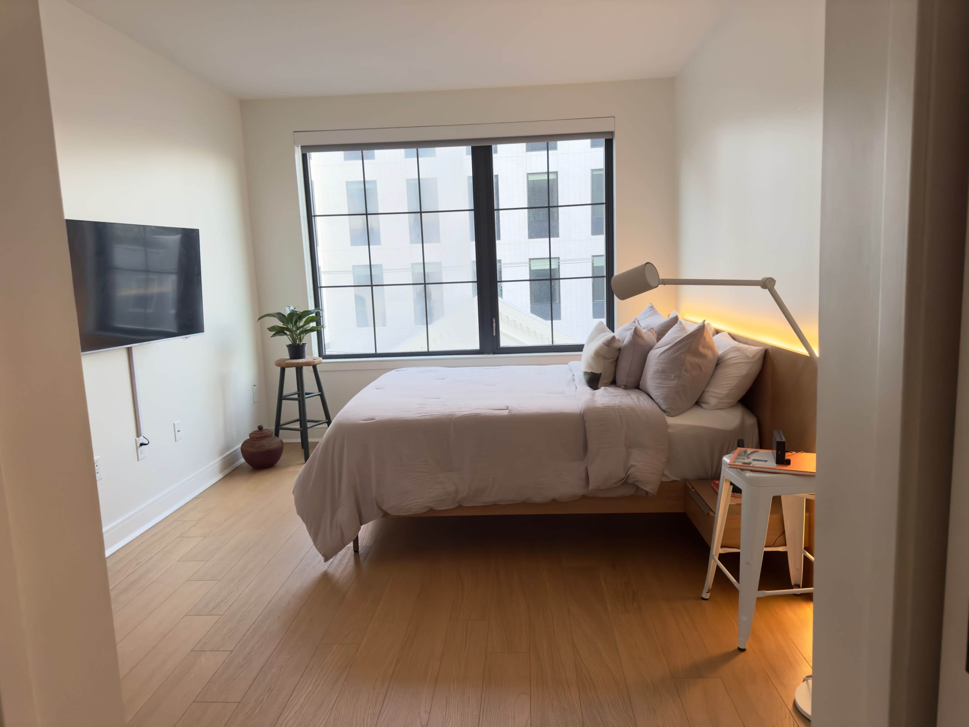 A bright bedroom features a bed with light bedding, a television mounted on the wall, and a potted plant beside a nightstand.