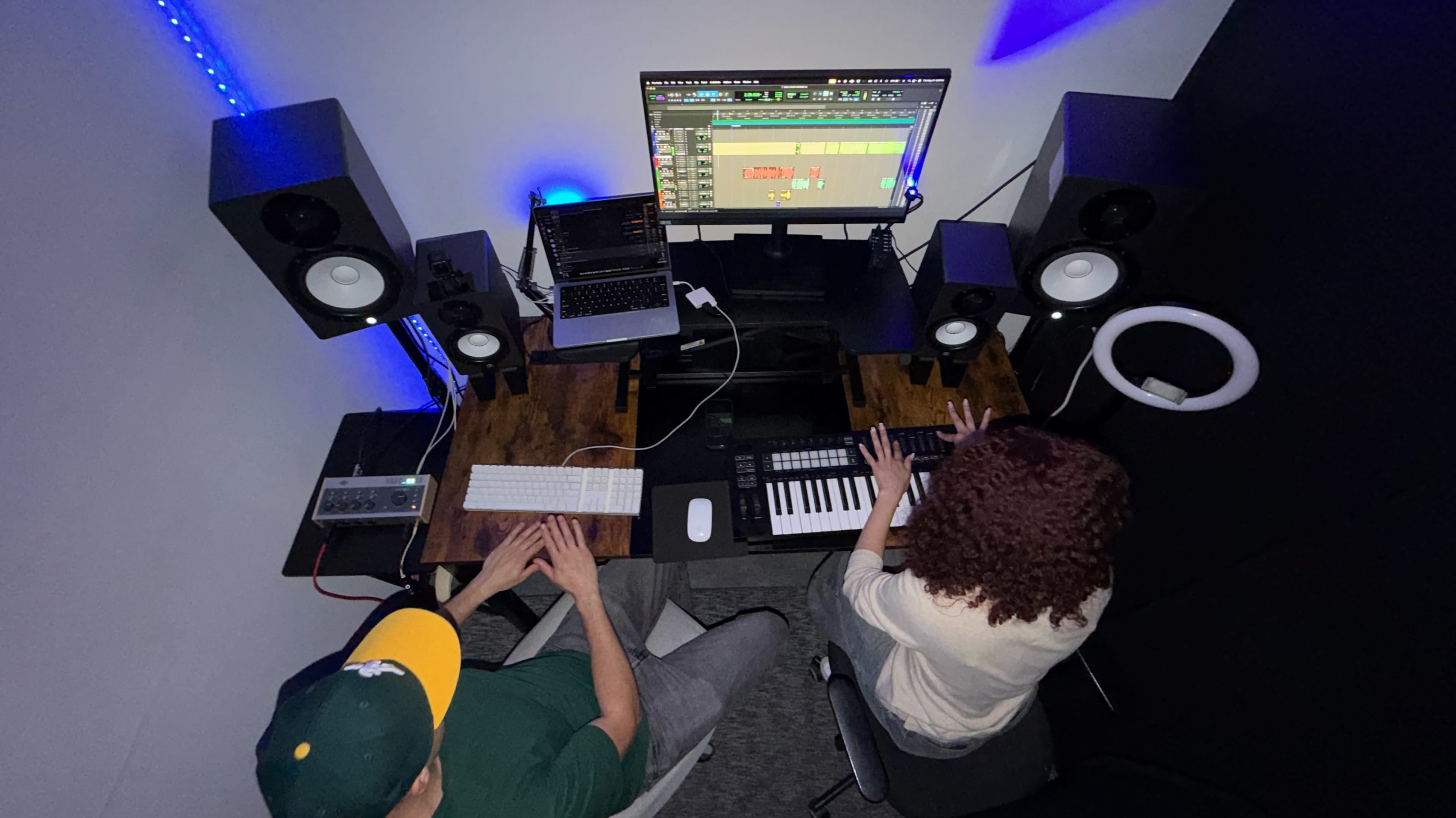 Two individuals are seated at a desk in a music production studio, working on a computer with a digital audio workstation open, surrounded by speakers and recording equipment.