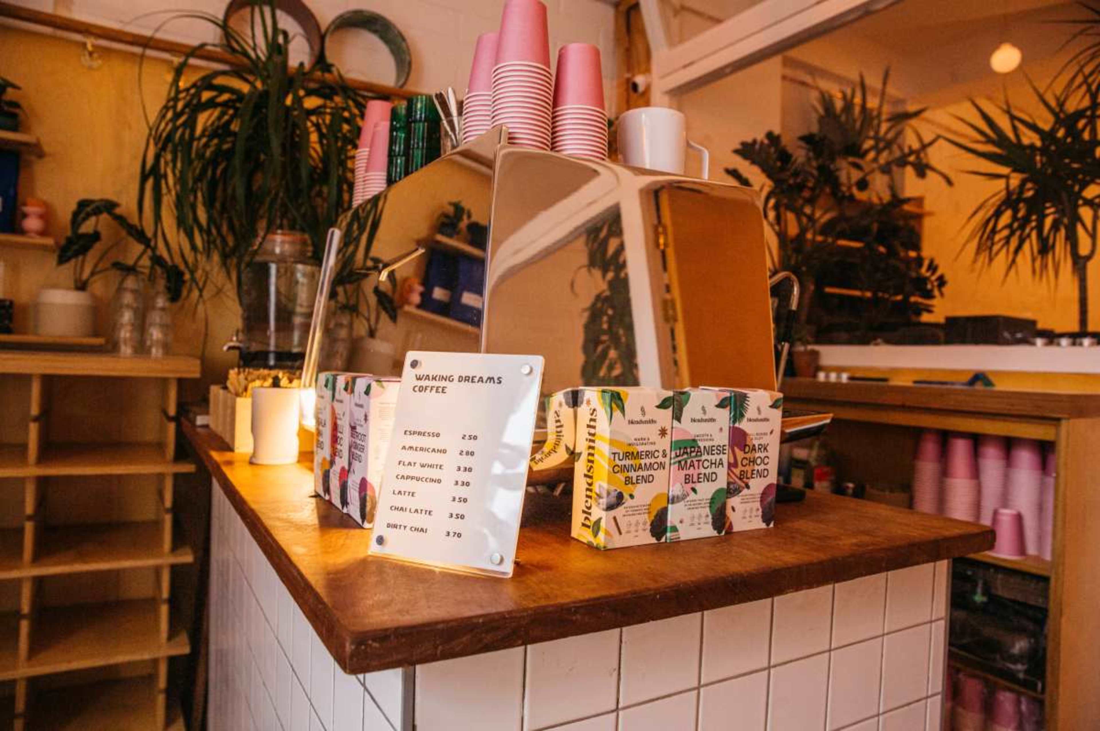 A coffee counter with a menu board and colorful packaging for coffee blends, surrounded by plants and paper cups.