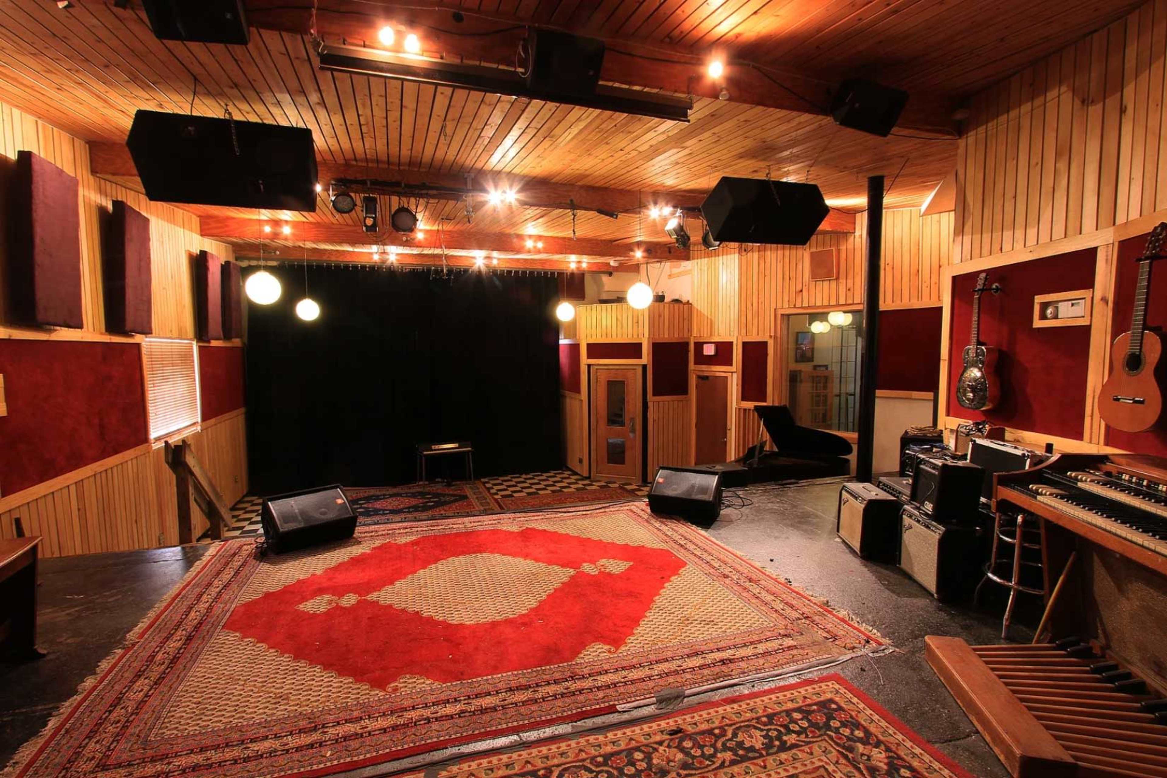 A music studio with a wooden ceiling, various musical instruments, and a stage area highlighted by overhead lights and a red rug.