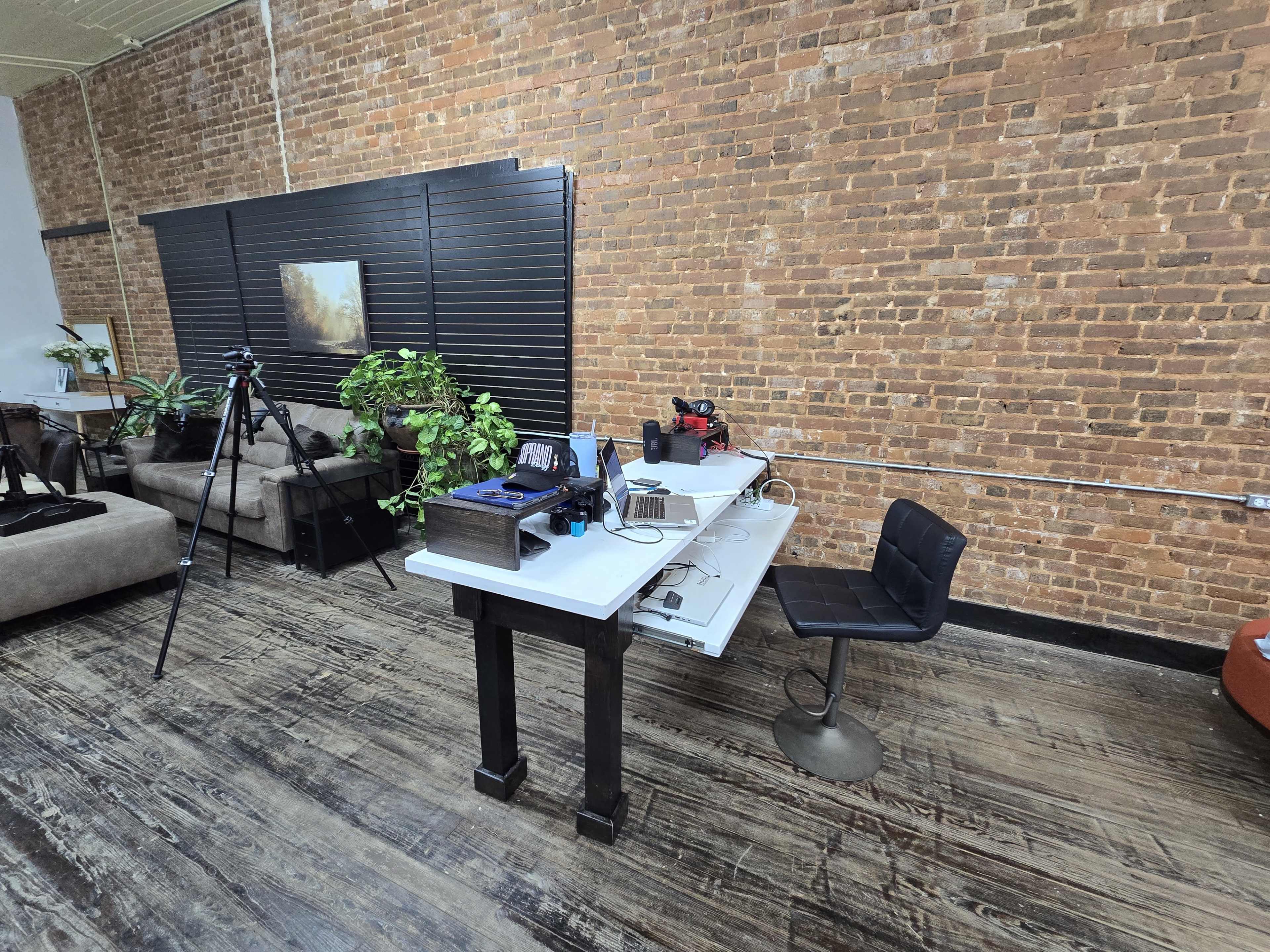 The image shows a workspace with a white table, a black chair, various tools, a camera setup on a tripod, and greenery against a brick wall backdrop.