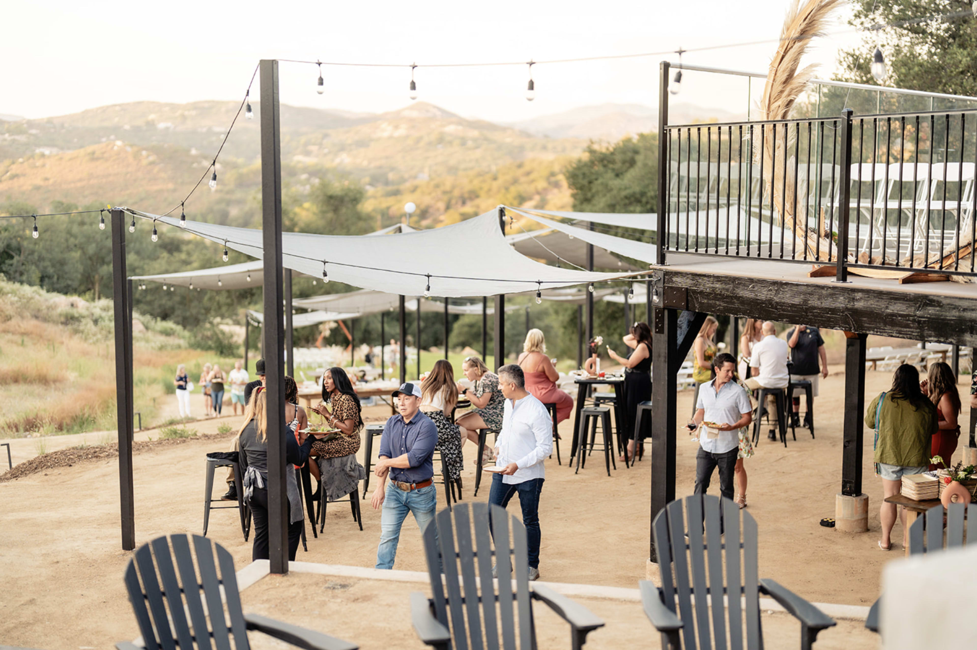 A group of people socializes at an outdoor event featuring string lights, shaded areas, and mountain views.