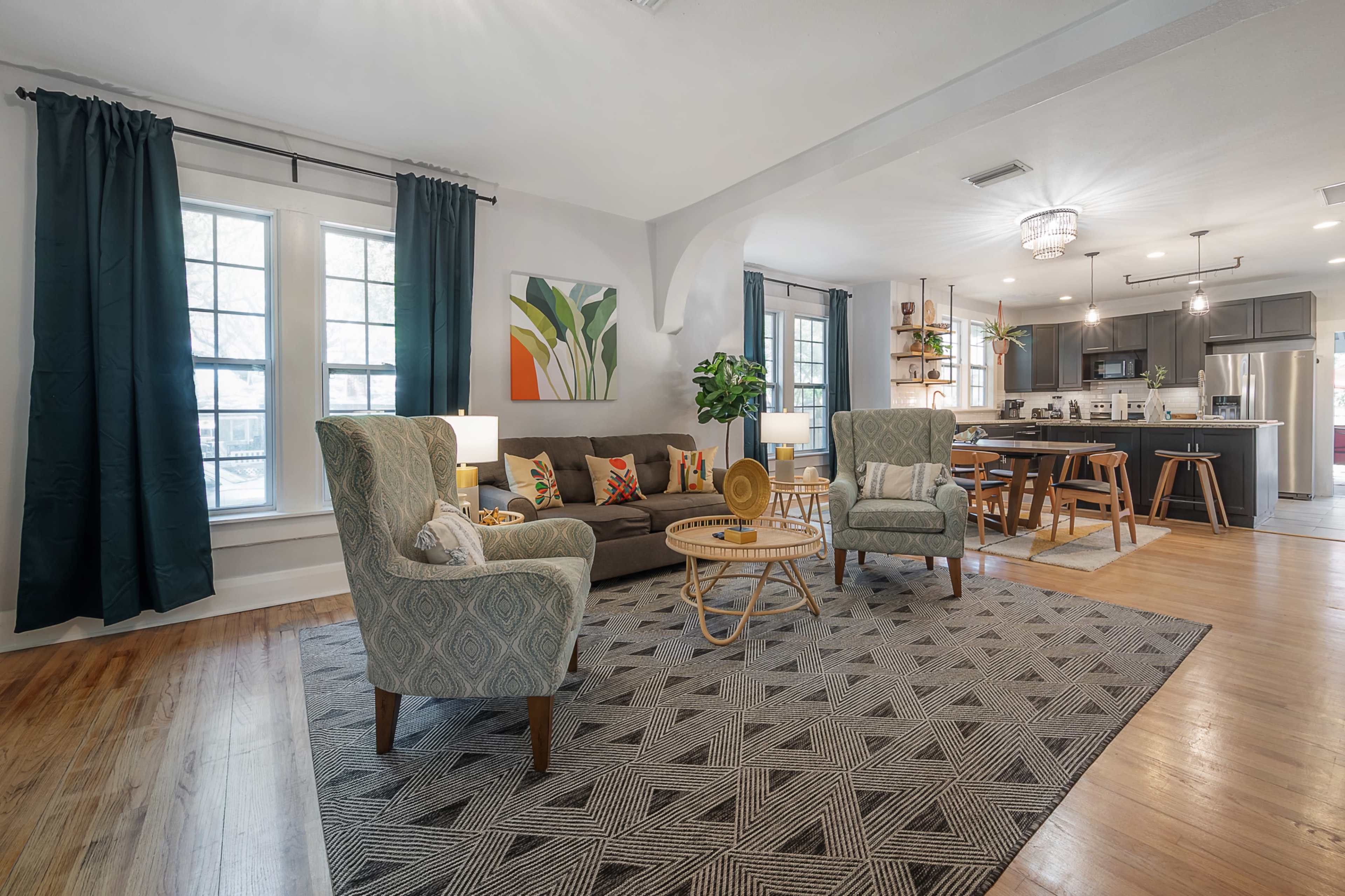 The living area features a gray sofa, two patterned chairs, a round coffee table, and a dining space with wooden chairs in a light-filled open layout.
