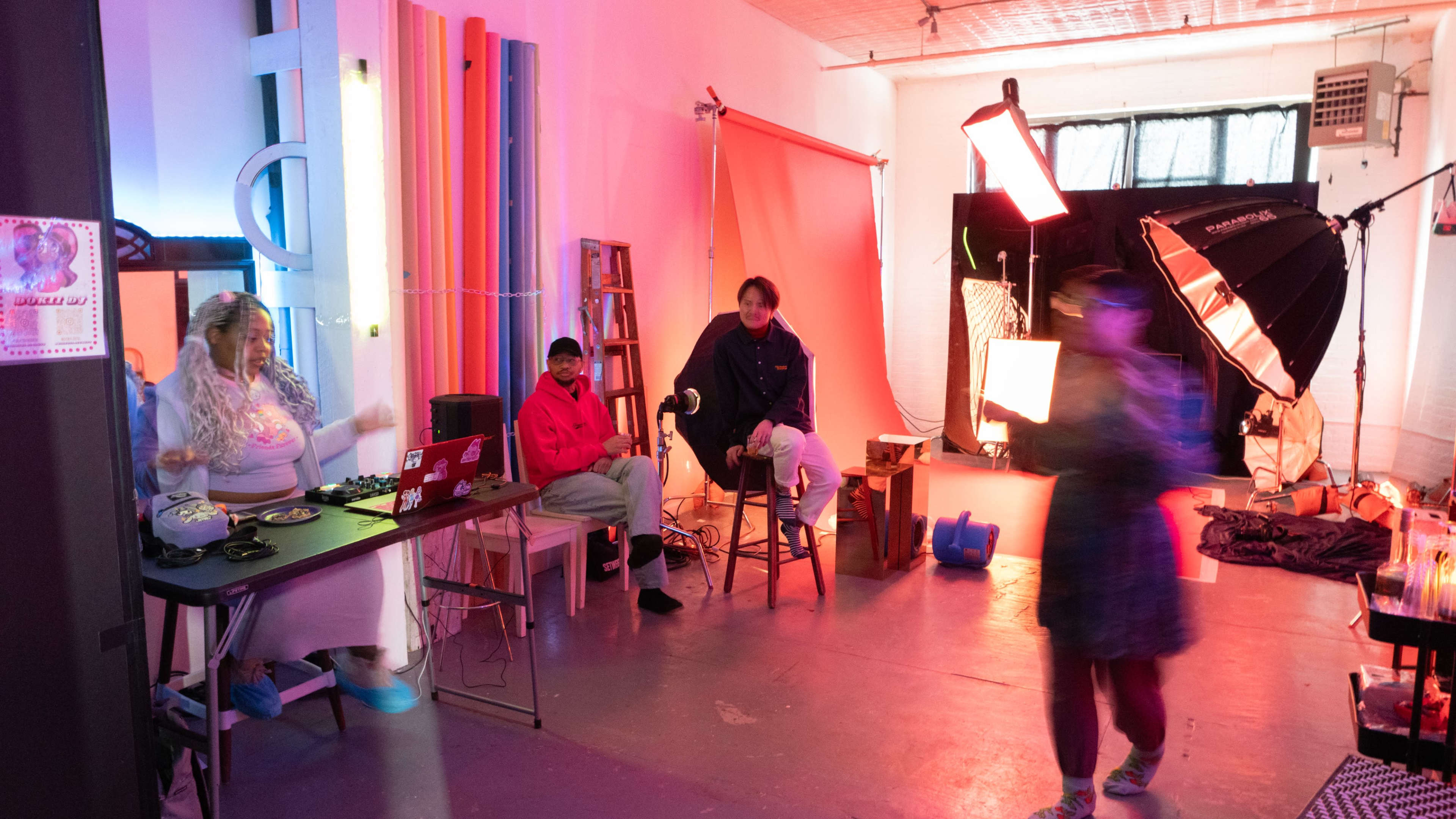 The image shows a studio space with colorful backdrops and lighting equipment, where two people are seated at a table while a third person stands in motion.