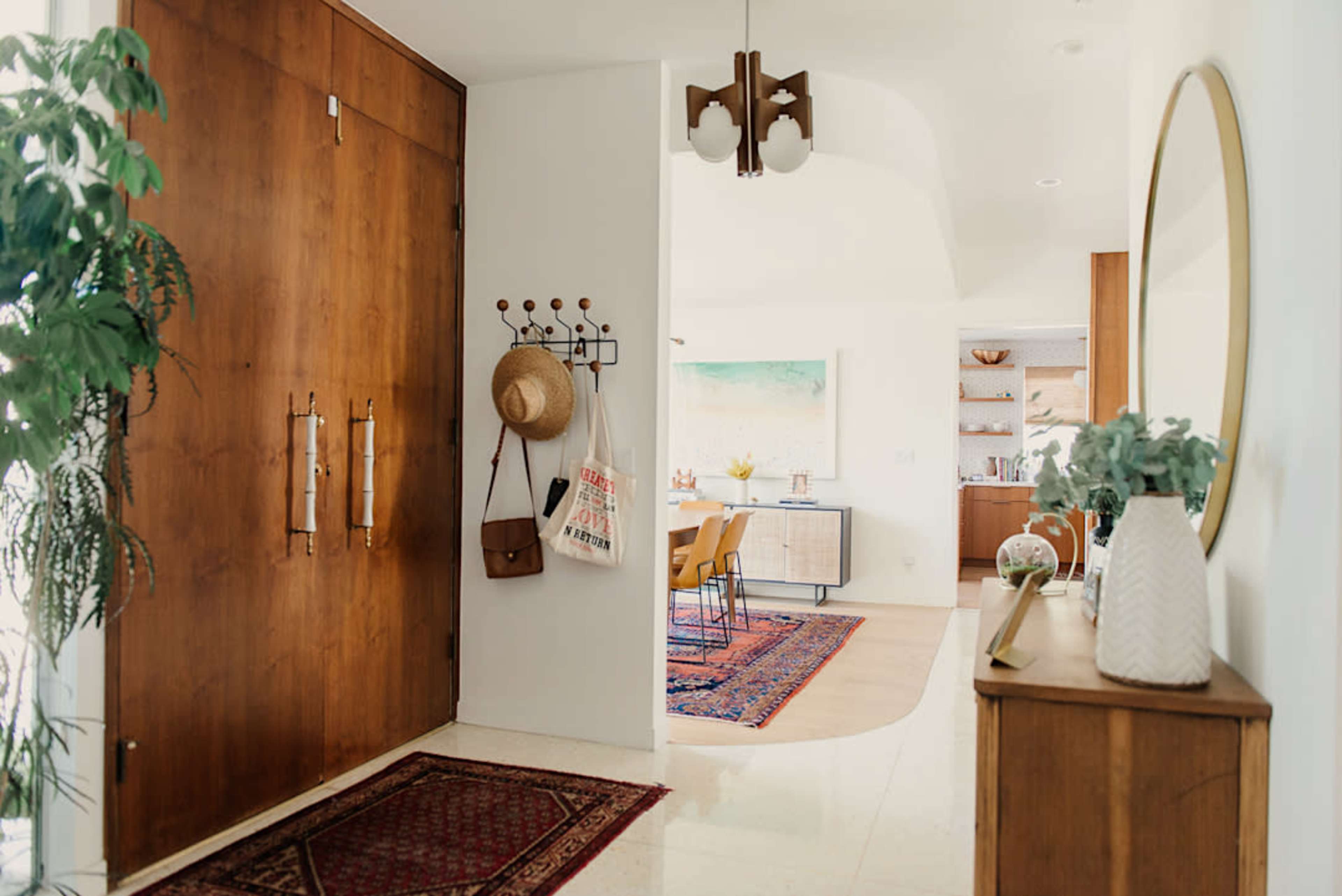 A spacious entryway features a large wooden door, a wall-mounted coat rack, and a circular mirror, leading to a bright dining area with a colorful rug.