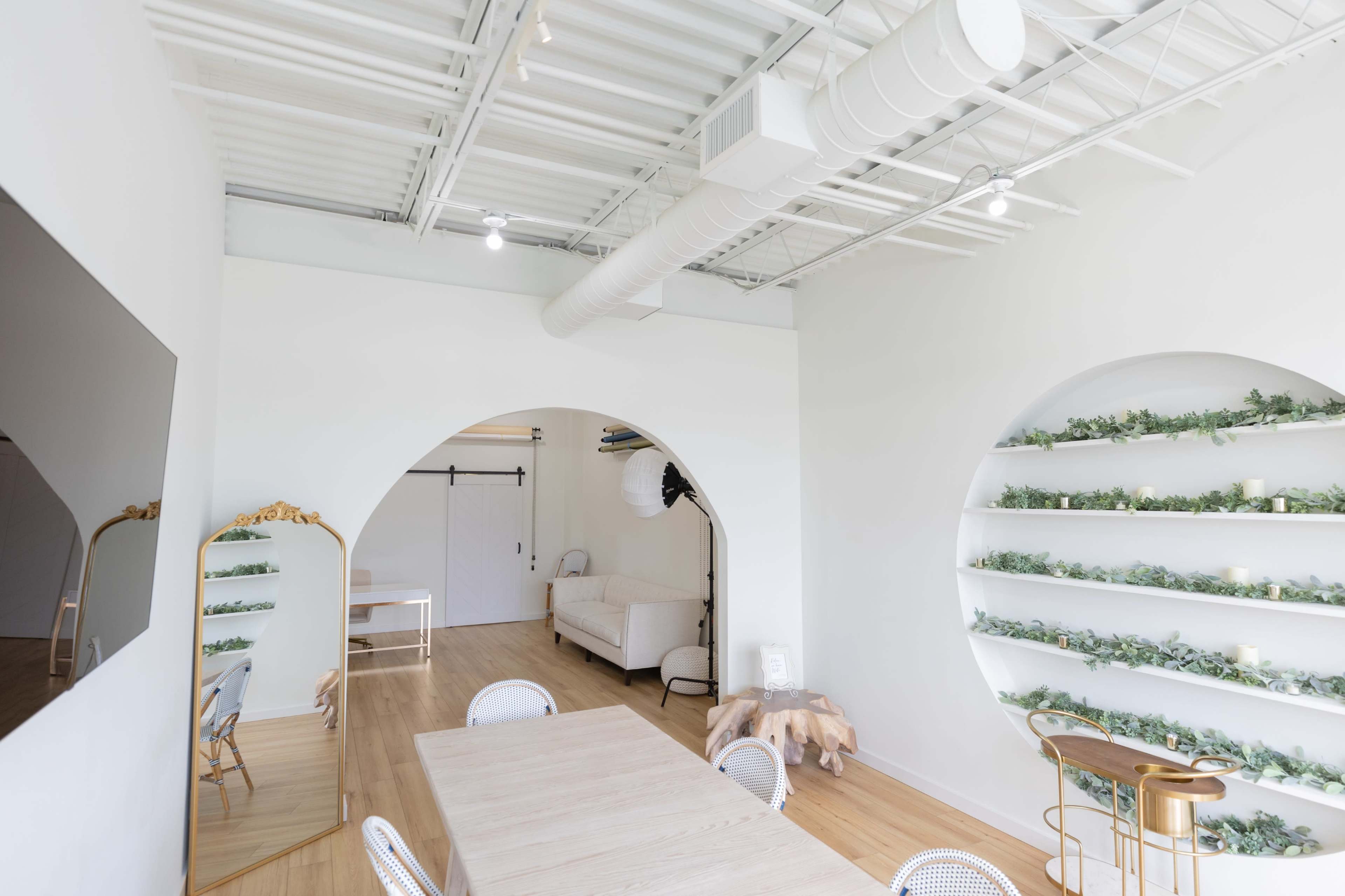 The image shows a bright, modern studio space featuring an arched wall with shelves adorned with greenery, a wooden dining table, and a large mirror.