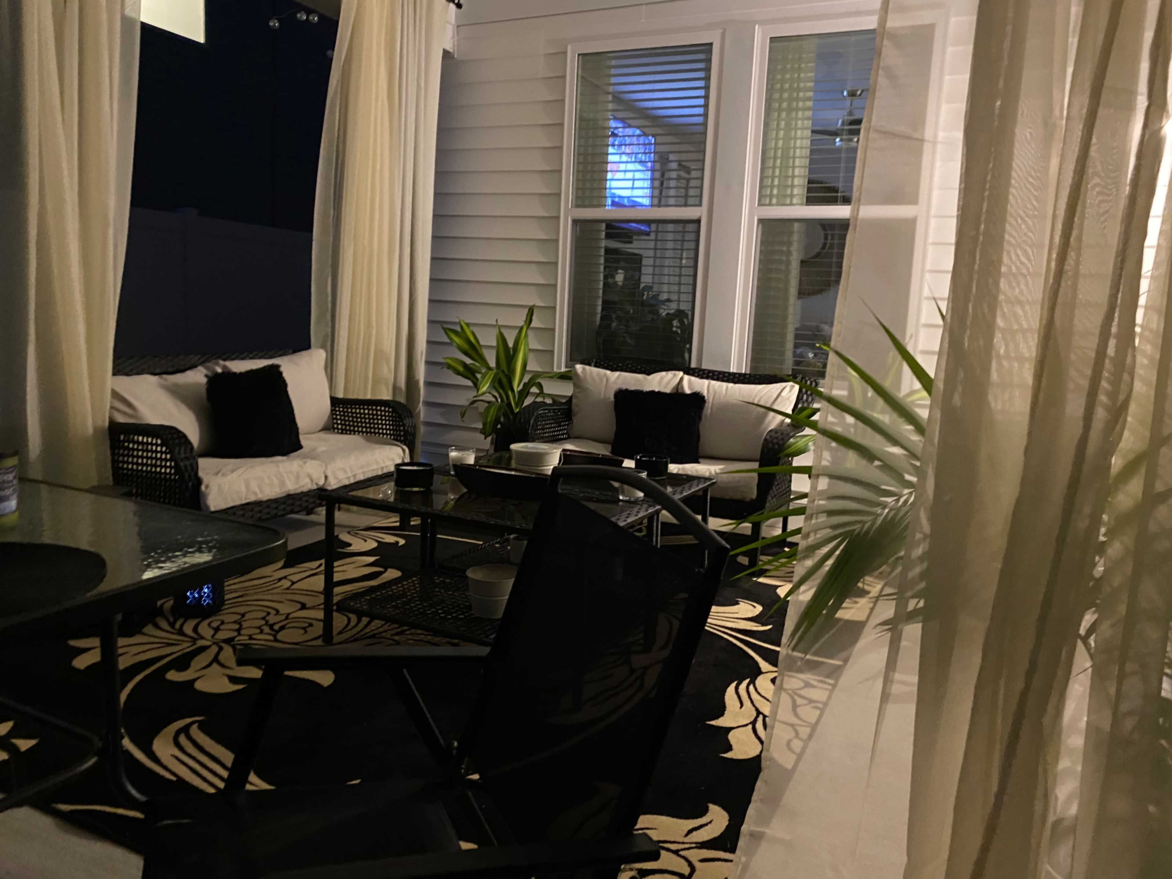 A cozy outdoor seating area with black wicker furniture, cushions, and planters, surrounded by light-colored curtains.