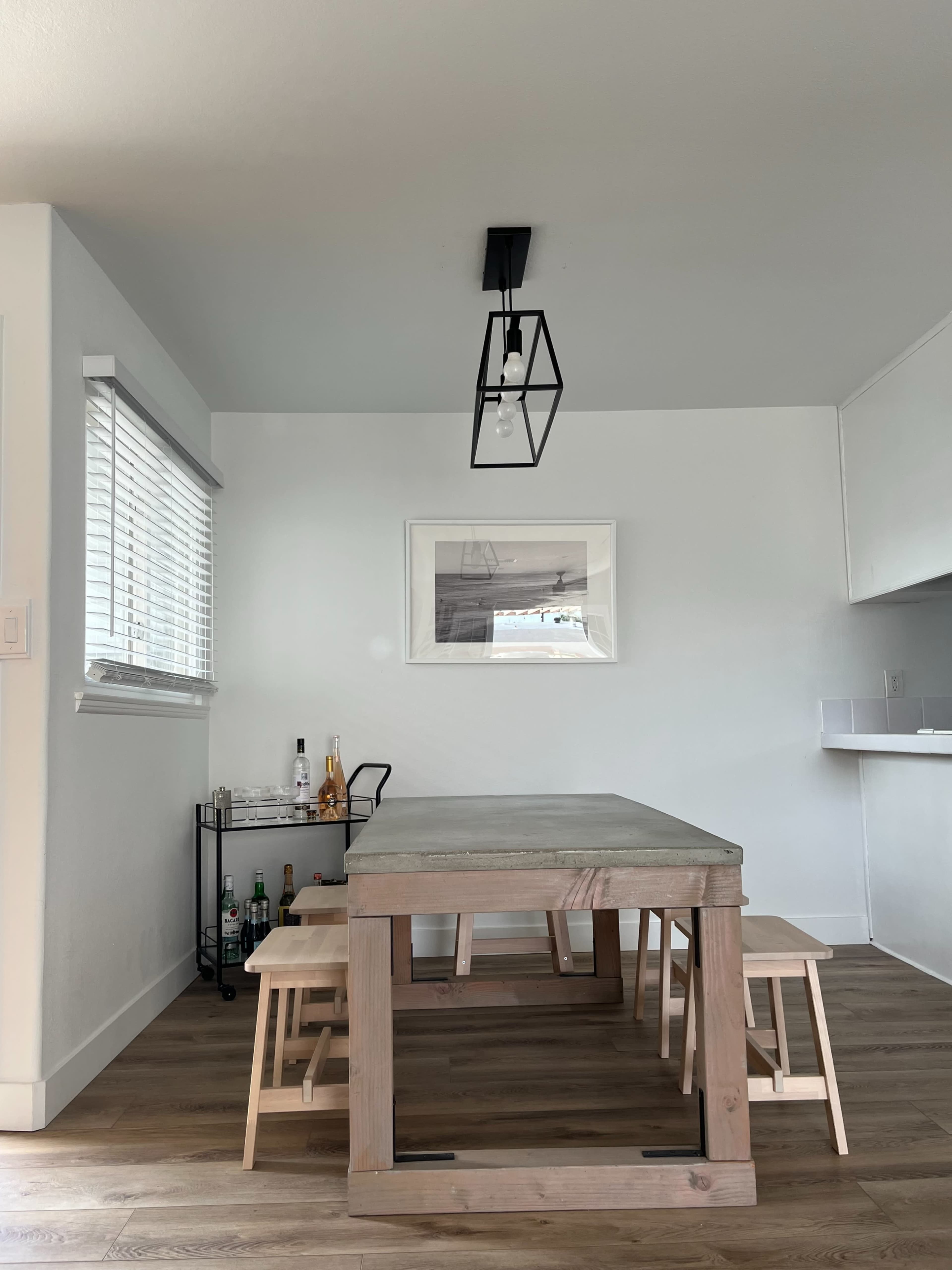 A minimalist dining area with a wooden table, two stools, a metal cart with bottles, and a framed photo on the white wall.
