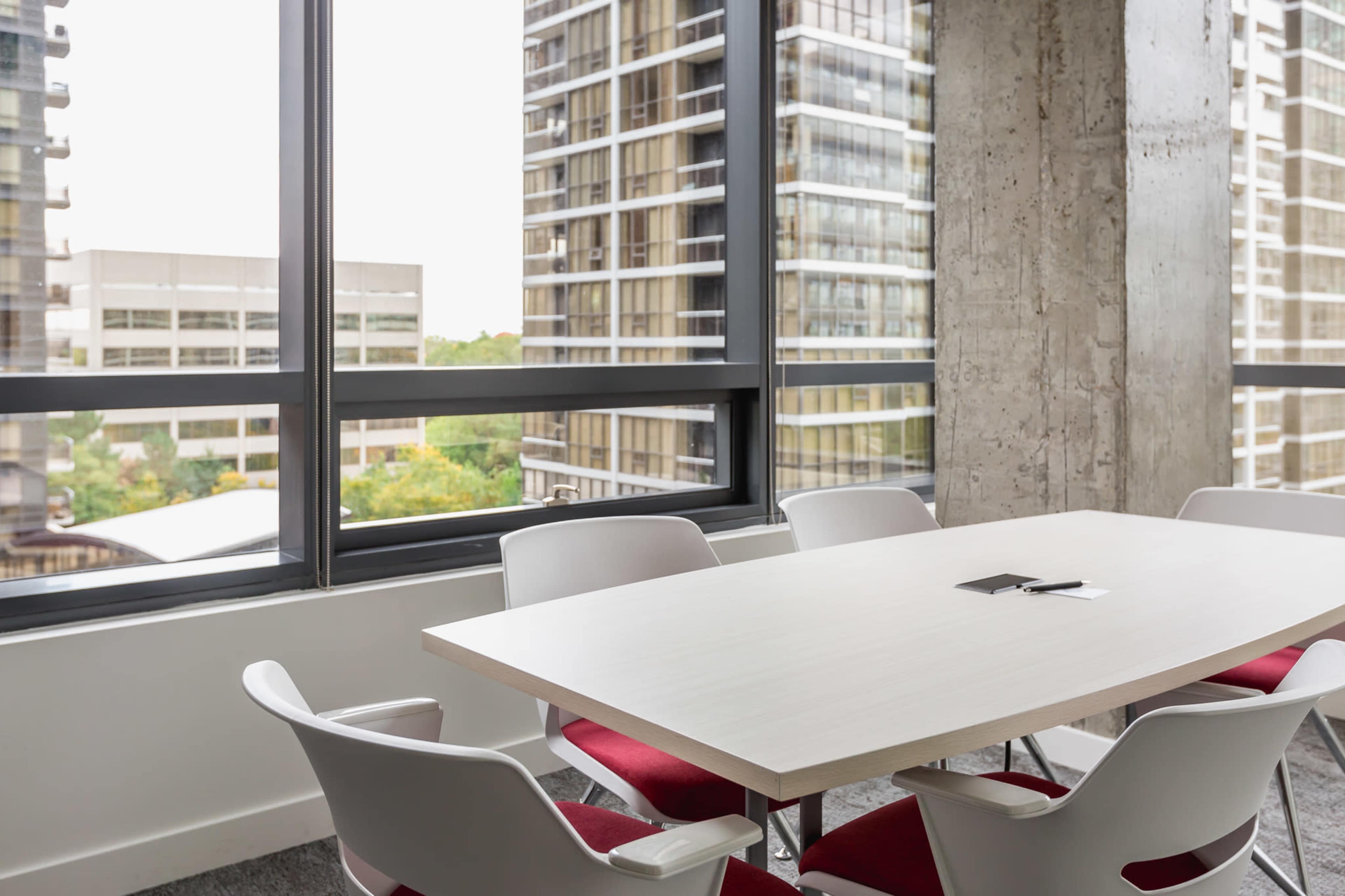 A modern conference room features a rectangular table surrounded by white chairs, with large windows displaying a view of nearby tall buildings.