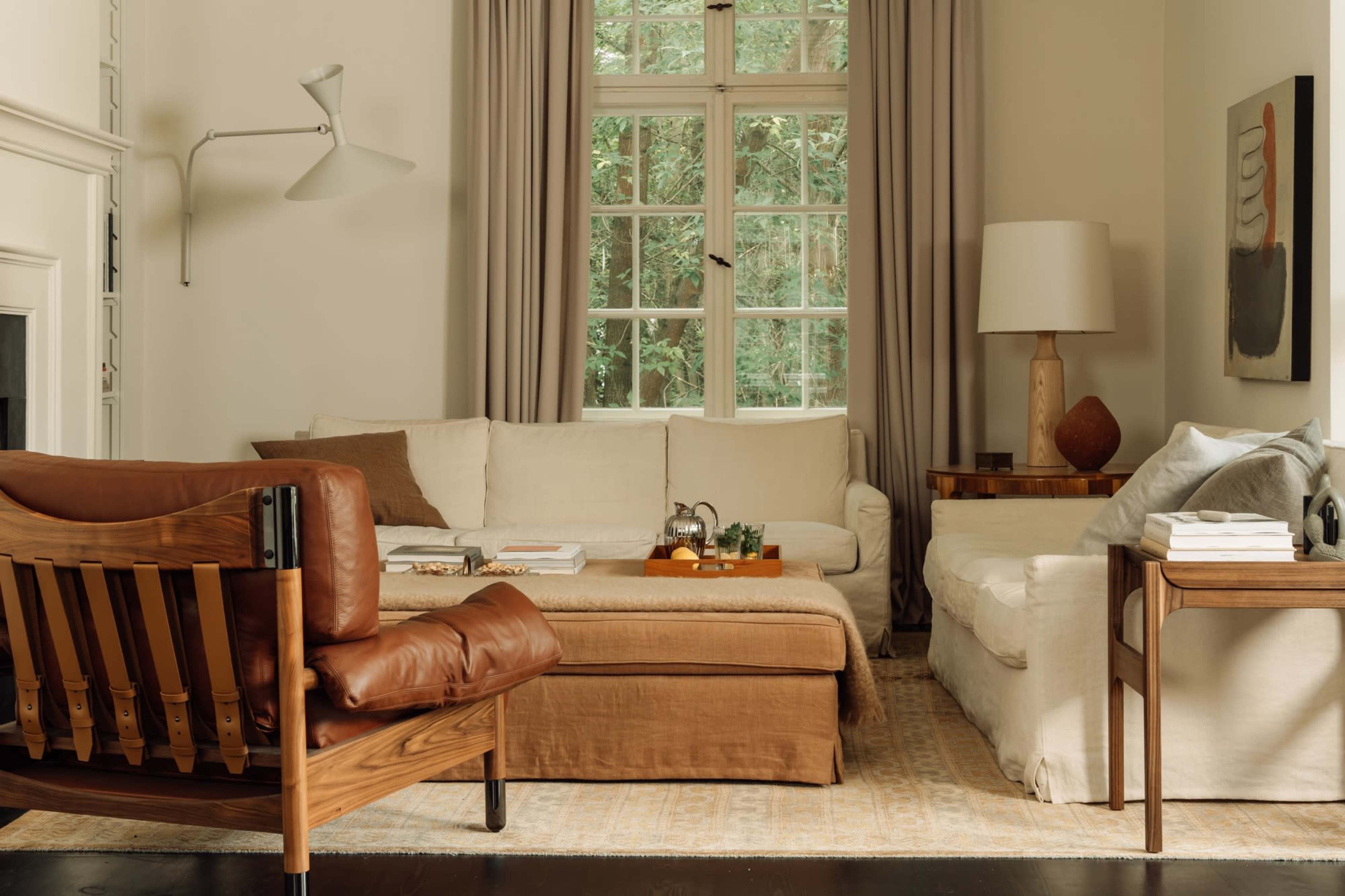 A cozy living room features a light-colored sofa, a dark wooden chair, and a coffee table surrounded by large windows with a view of greenery.