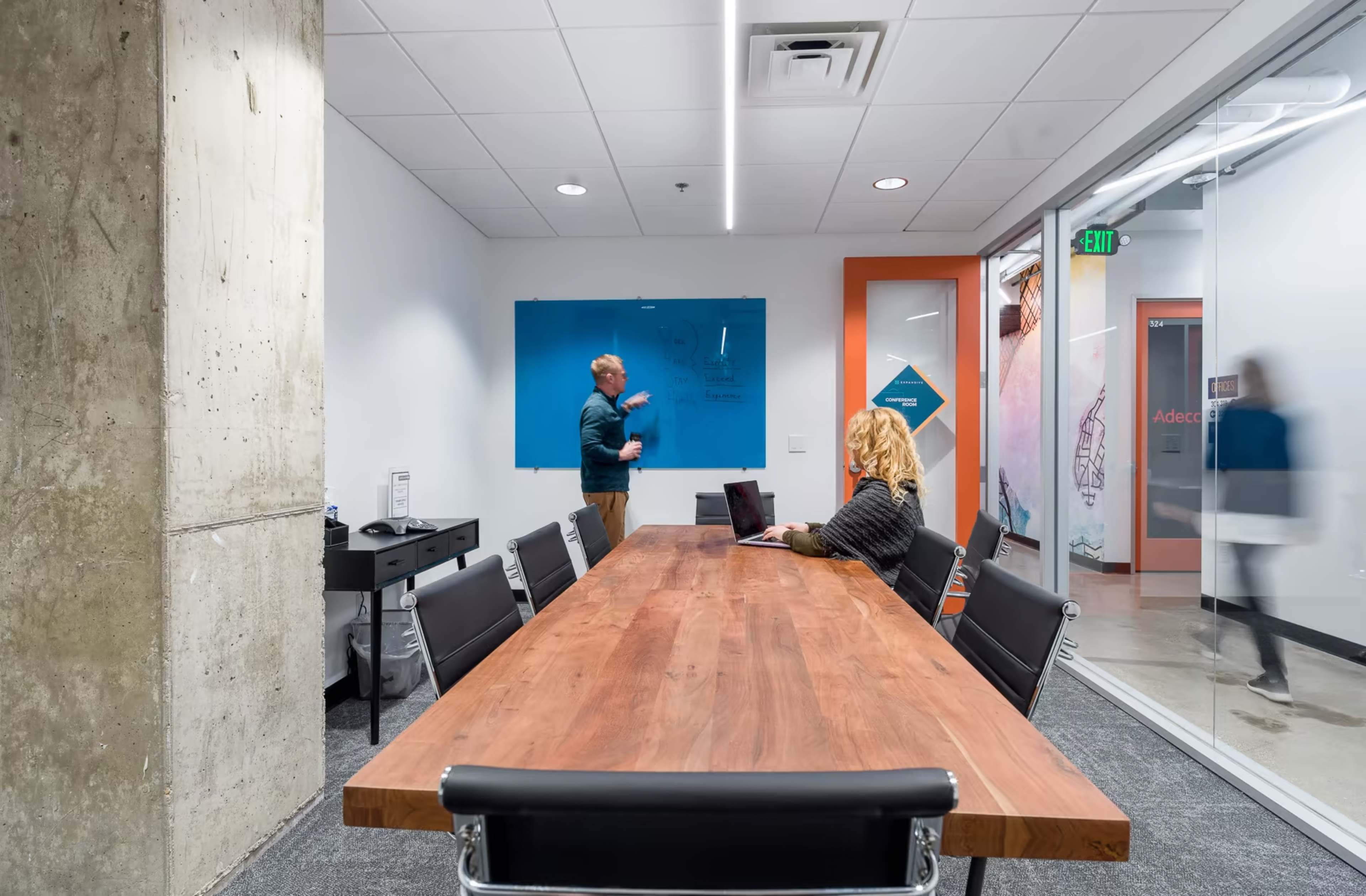 A man writes on a blue whiteboard while a woman sits at a wooden conference table with a laptop in a modern office meeting room.