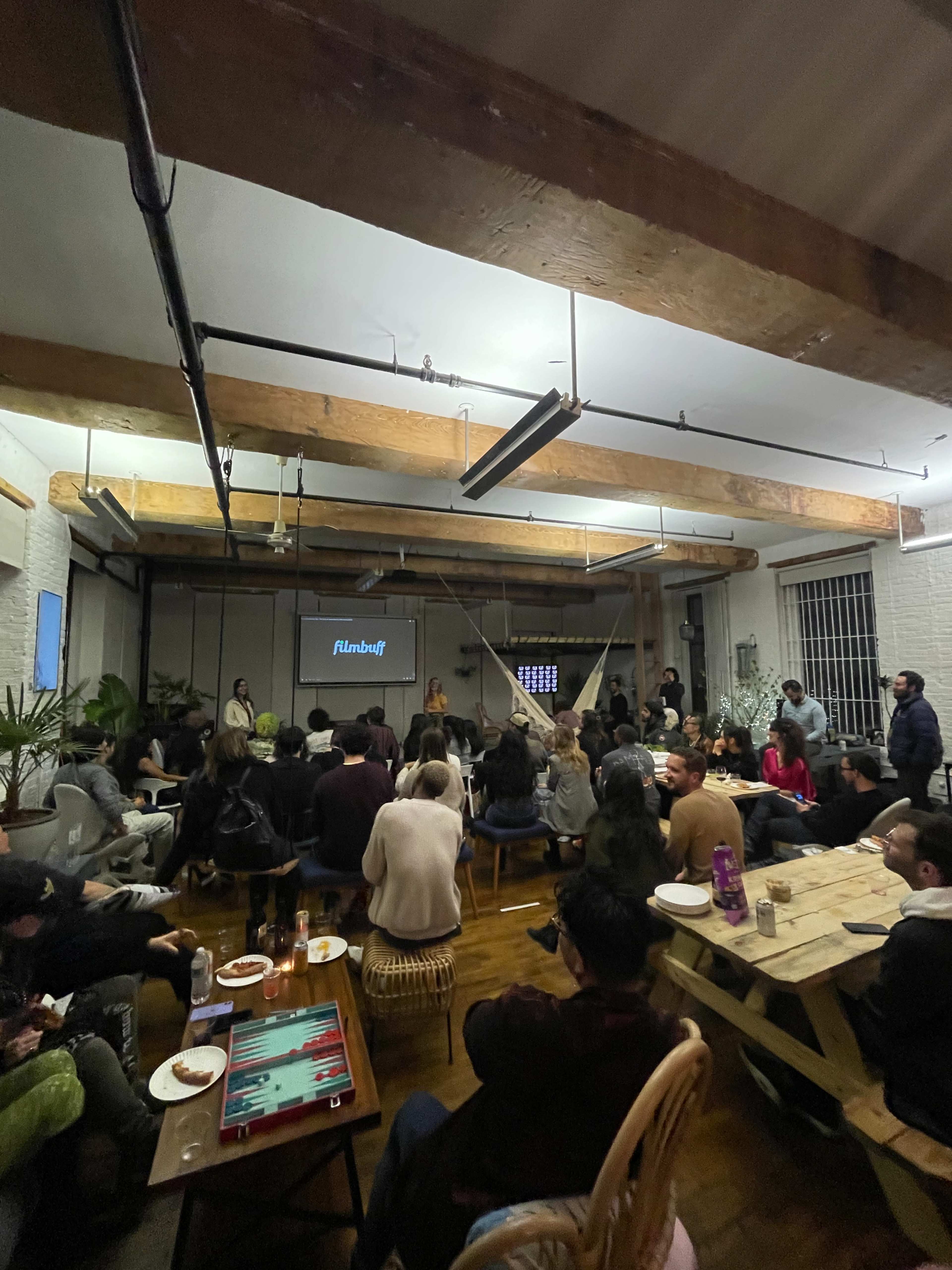 A group of people is seated in a spacious room with wooden beams, watching a presentation on a screen while food and drinks are placed on tables.