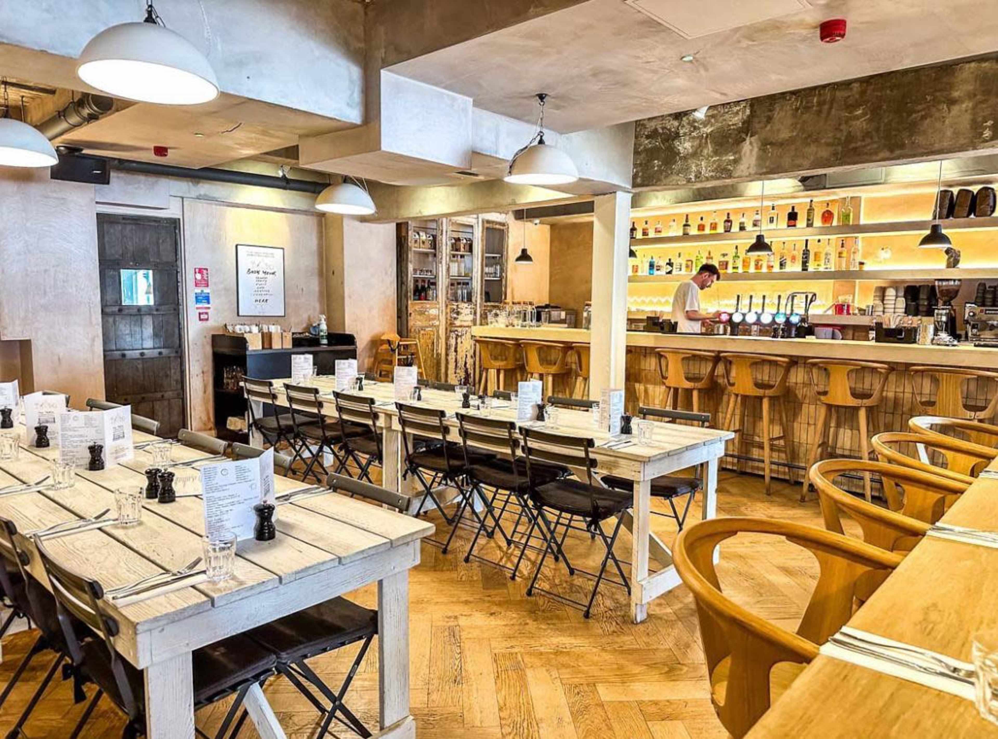 A modern restaurant interior with wooden tables, black chairs, and a bar featuring shelves of bottles in the background.