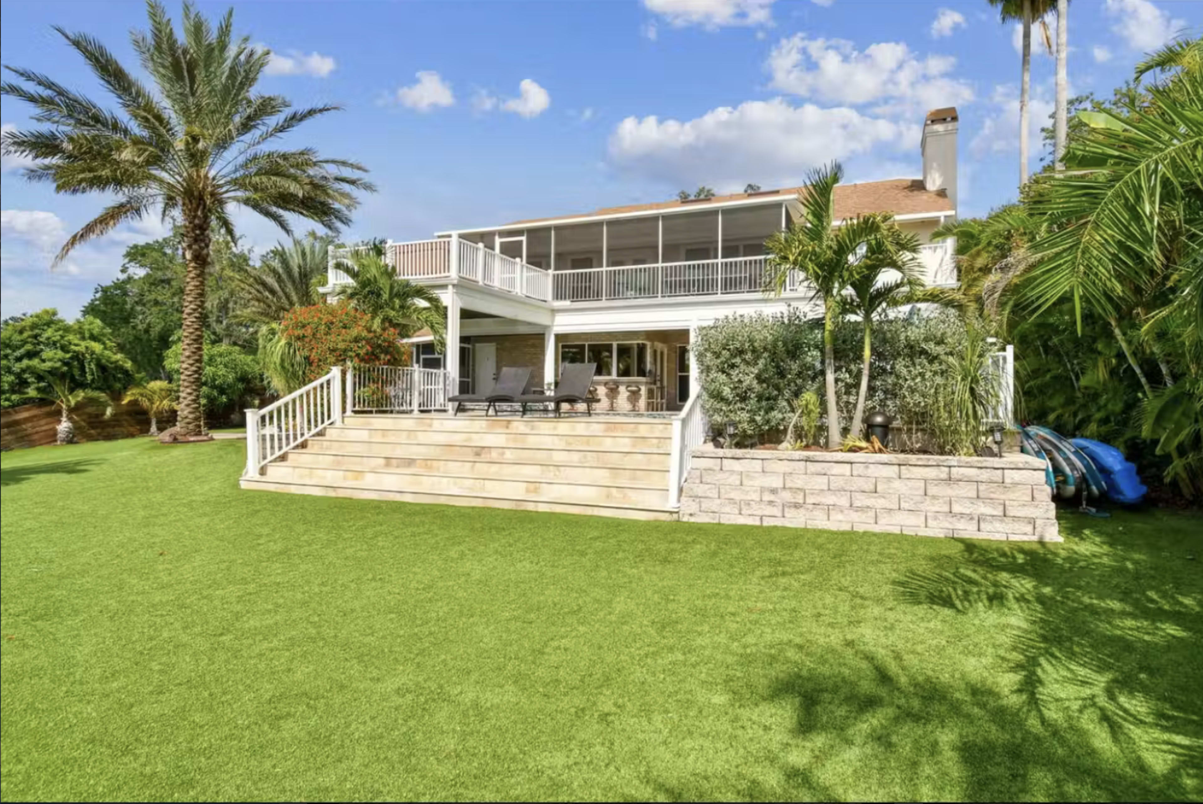 The image shows a two-story home with a spacious backyard featuring green grass, palm trees, and a patio area with outdoor furniture.