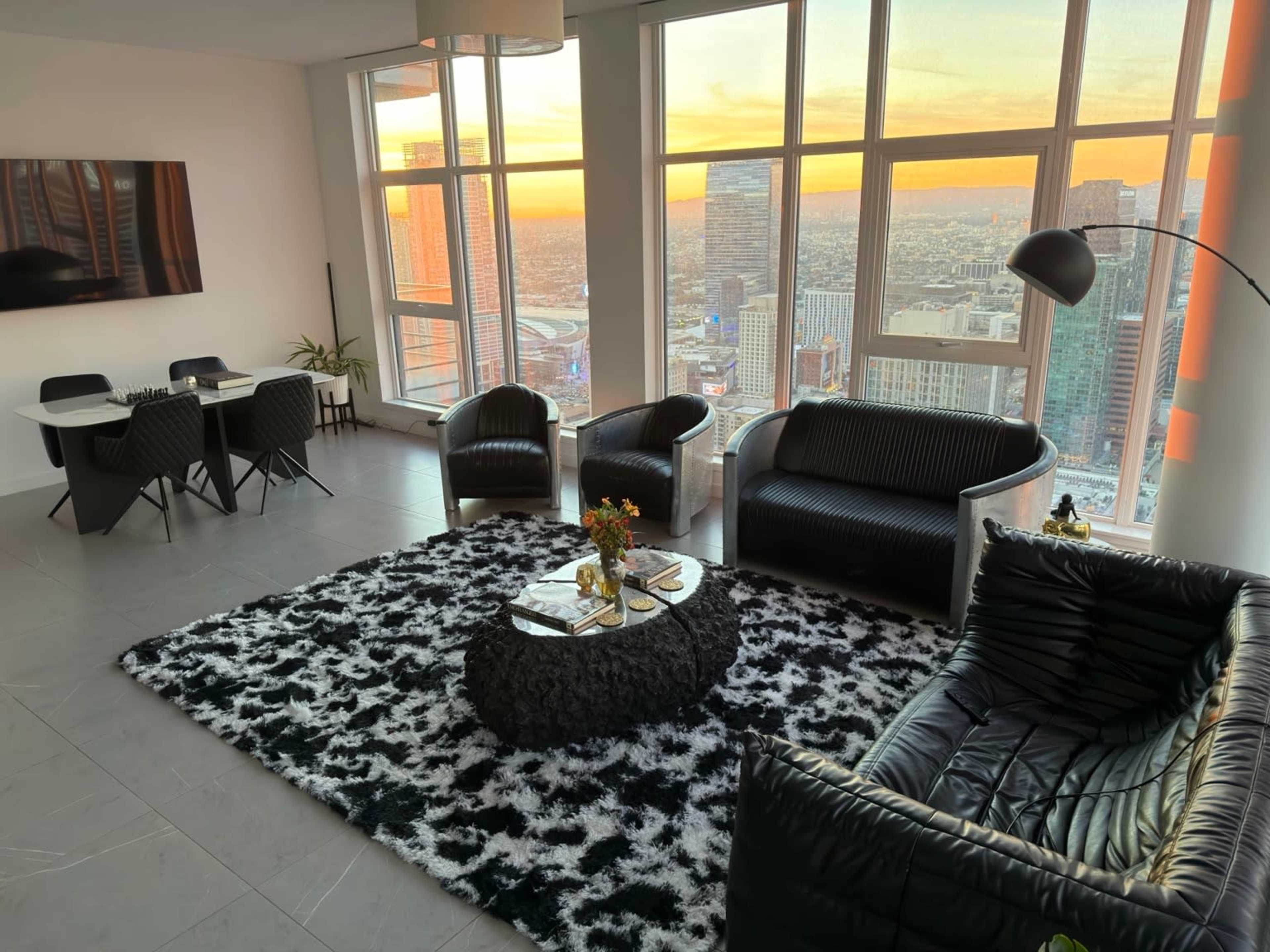 The image shows a modern living room with large windows overlooking a city skyline at sunset, featuring black leather furniture, a plush rug, and a coffee table.