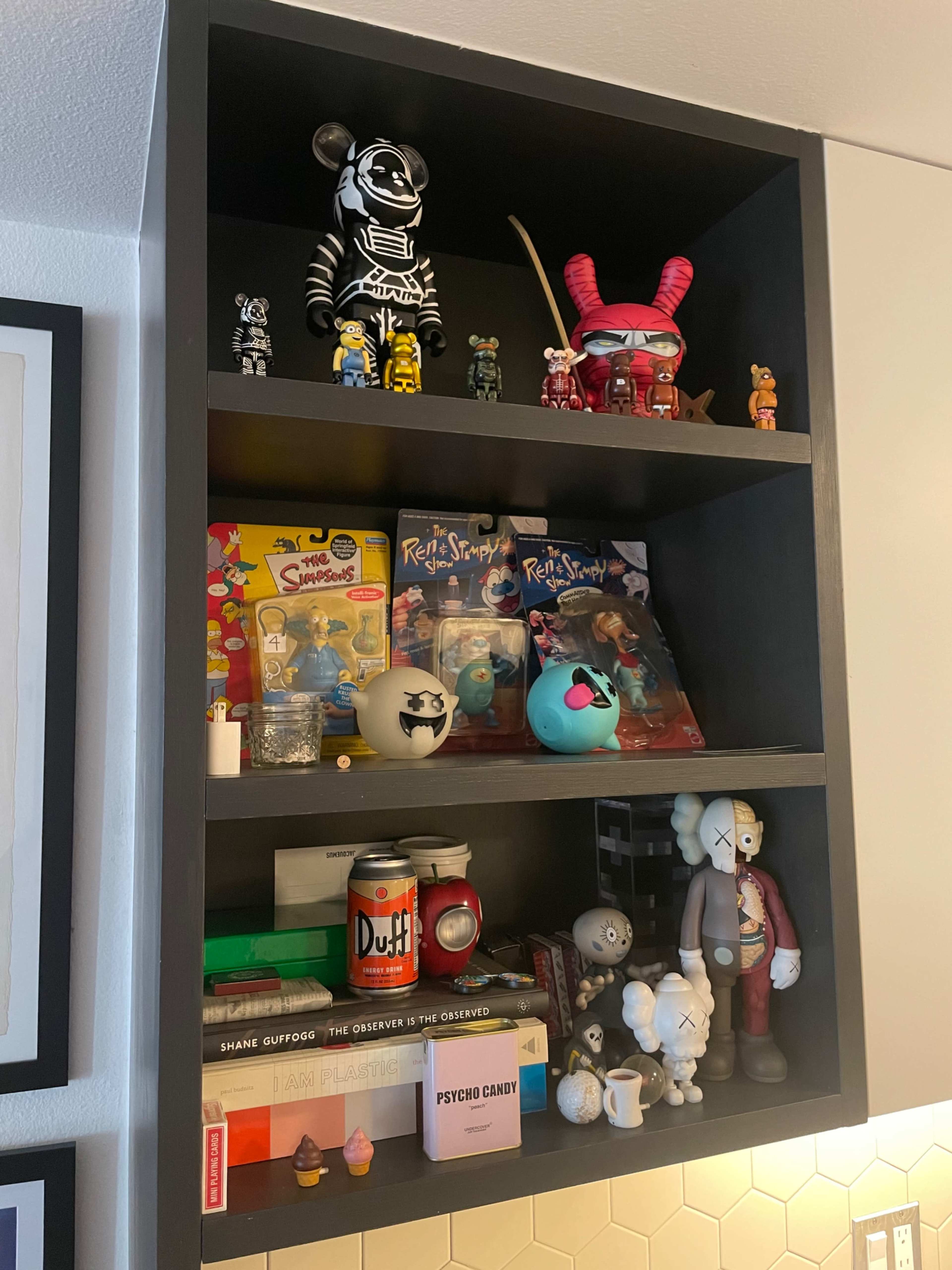 A black shelving unit displays various toys, collectibles, snacks, and books arranged across three shelves.