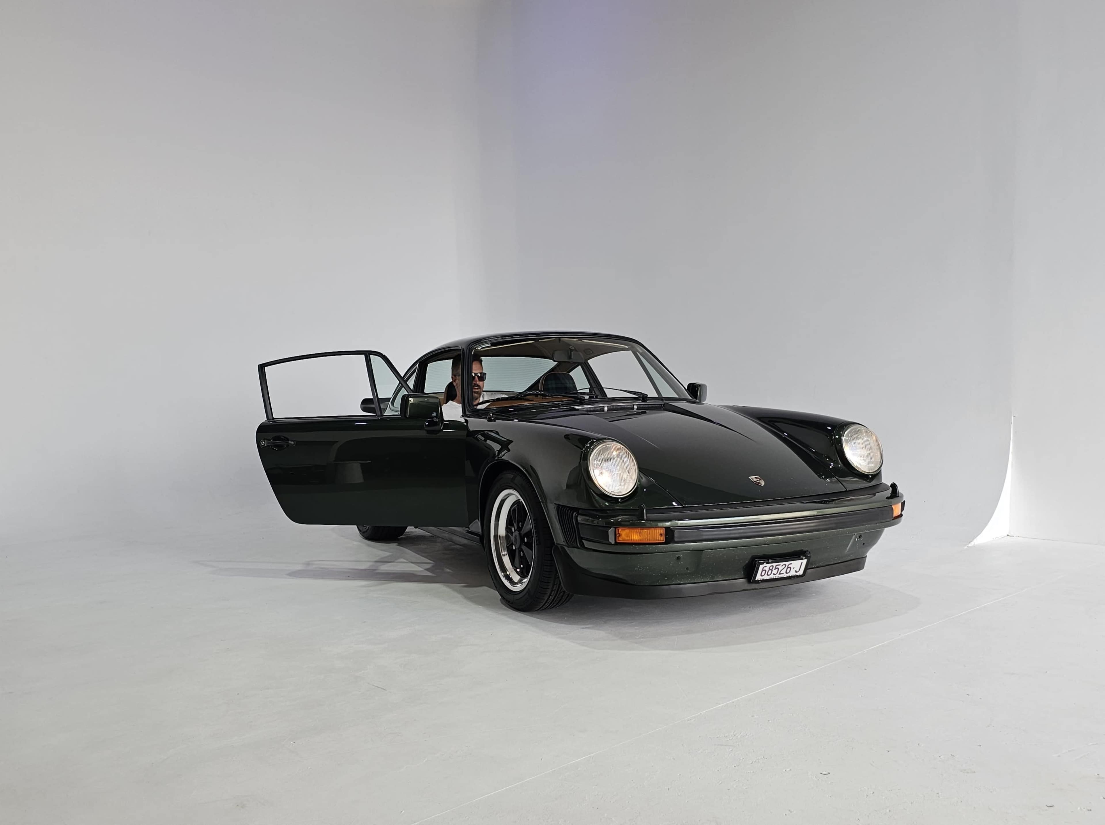 A black Porsche 911 is parked in an empty studio space with its driver's door open.