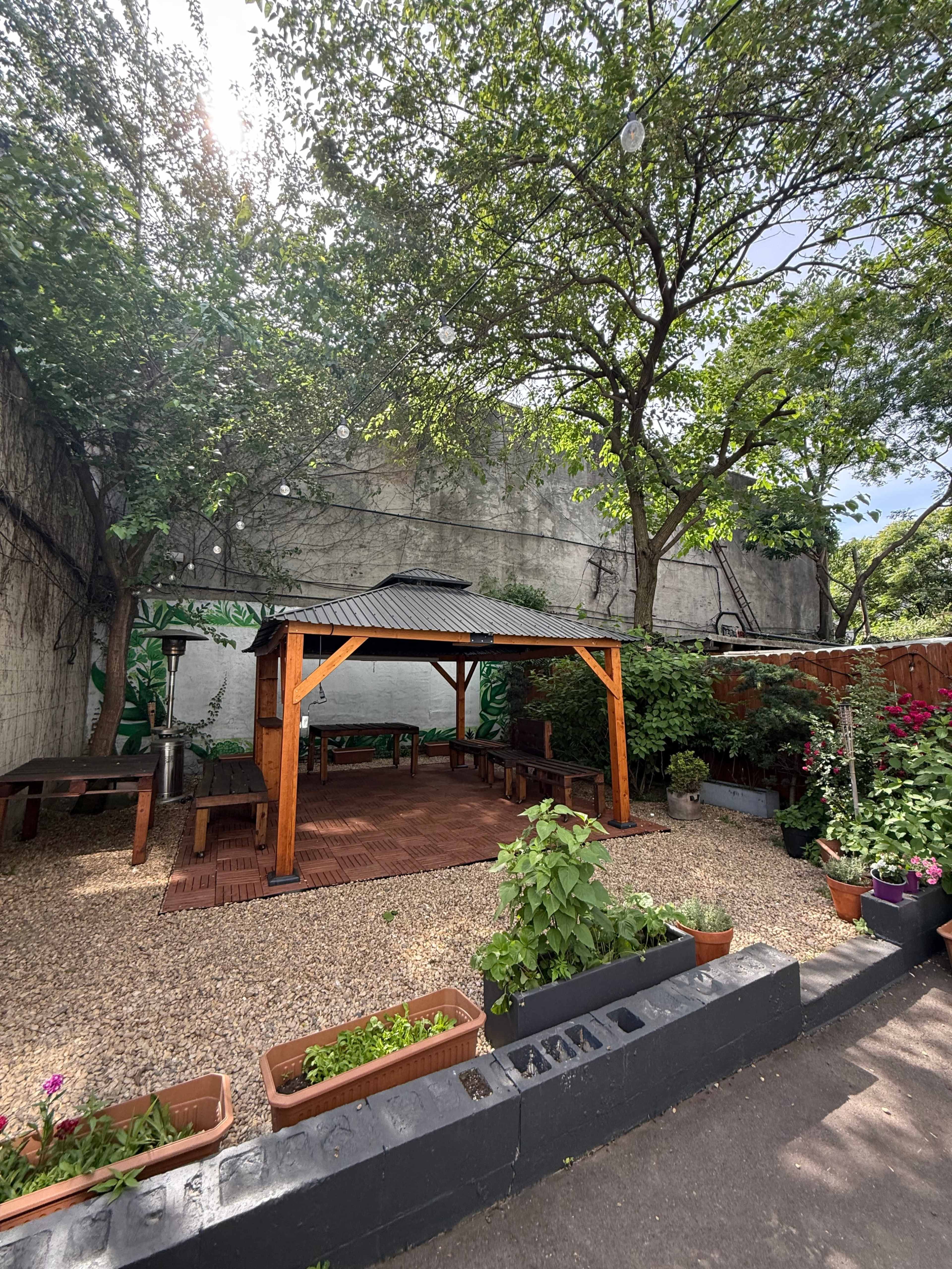The image shows a garden area with a wooden gazebo surrounded by potted plants and gravel pathways, beneath trees and a wall.