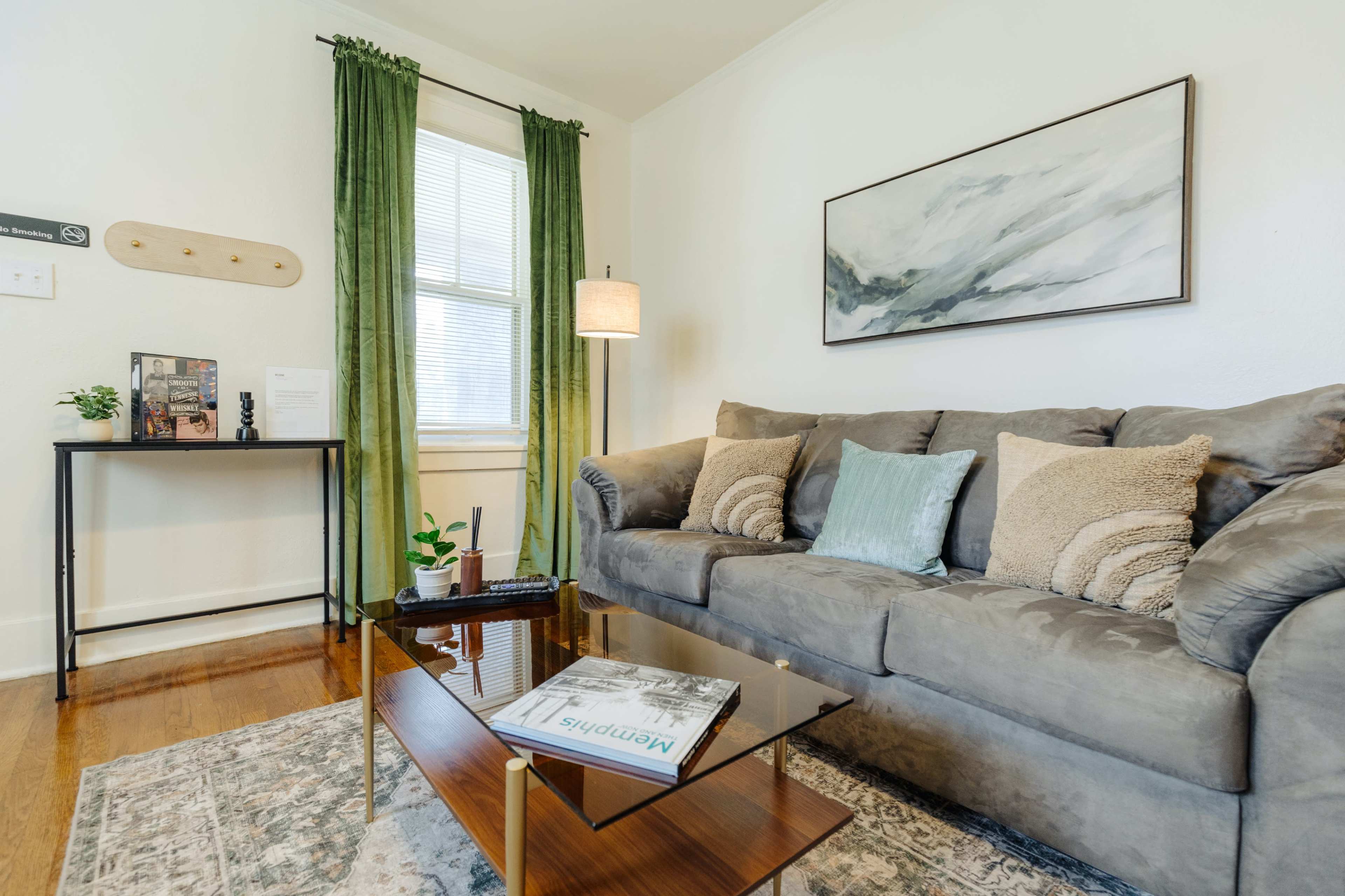 A modern living room features a gray sofa adorned with pillows, a glass coffee table, and a small black side table, all in front of a window with green curtains.