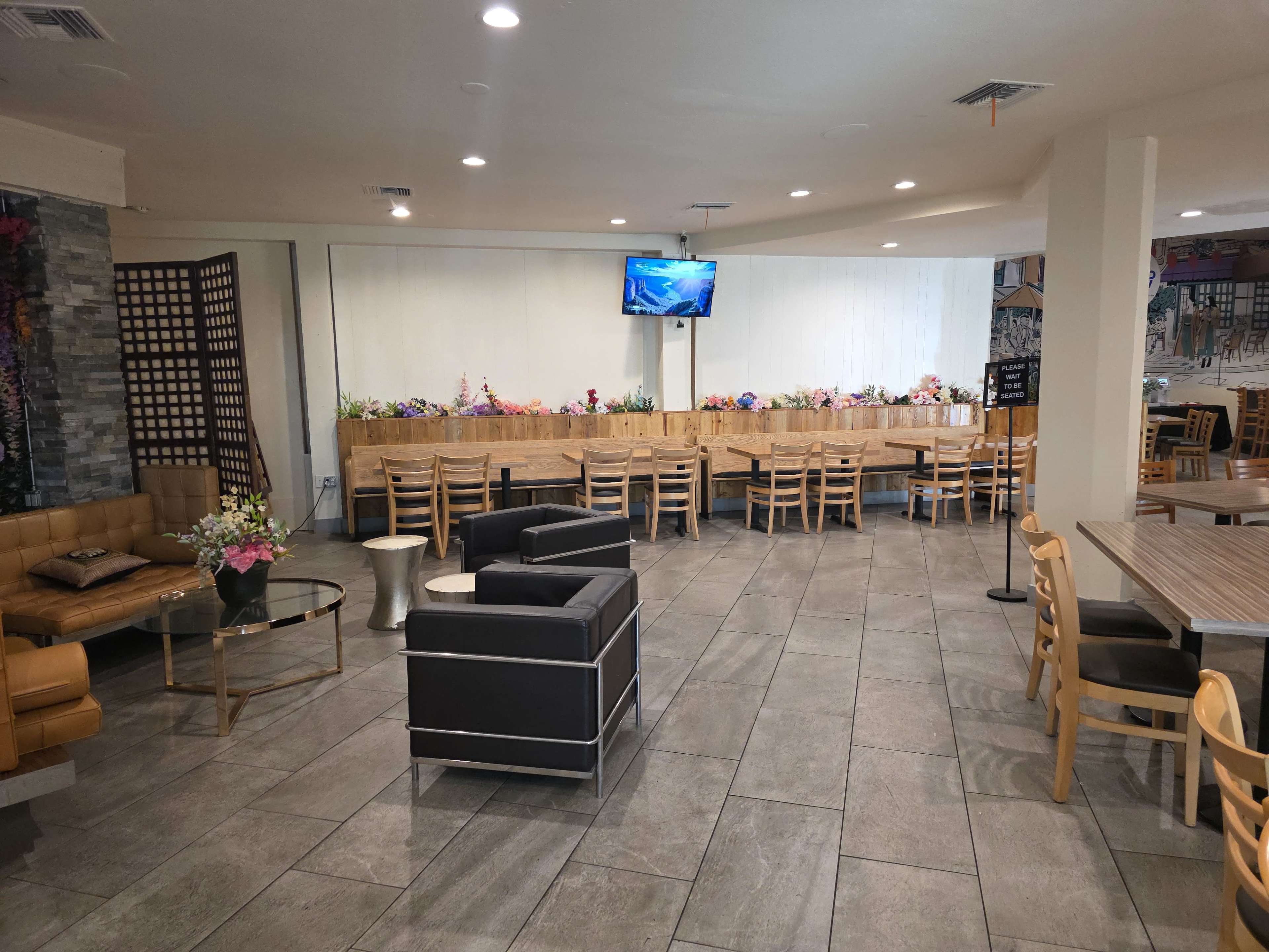 The interior of a restaurant features a seating area with wooden tables and chairs, a long bench adorned with flowers, and a television mounted on the wall.