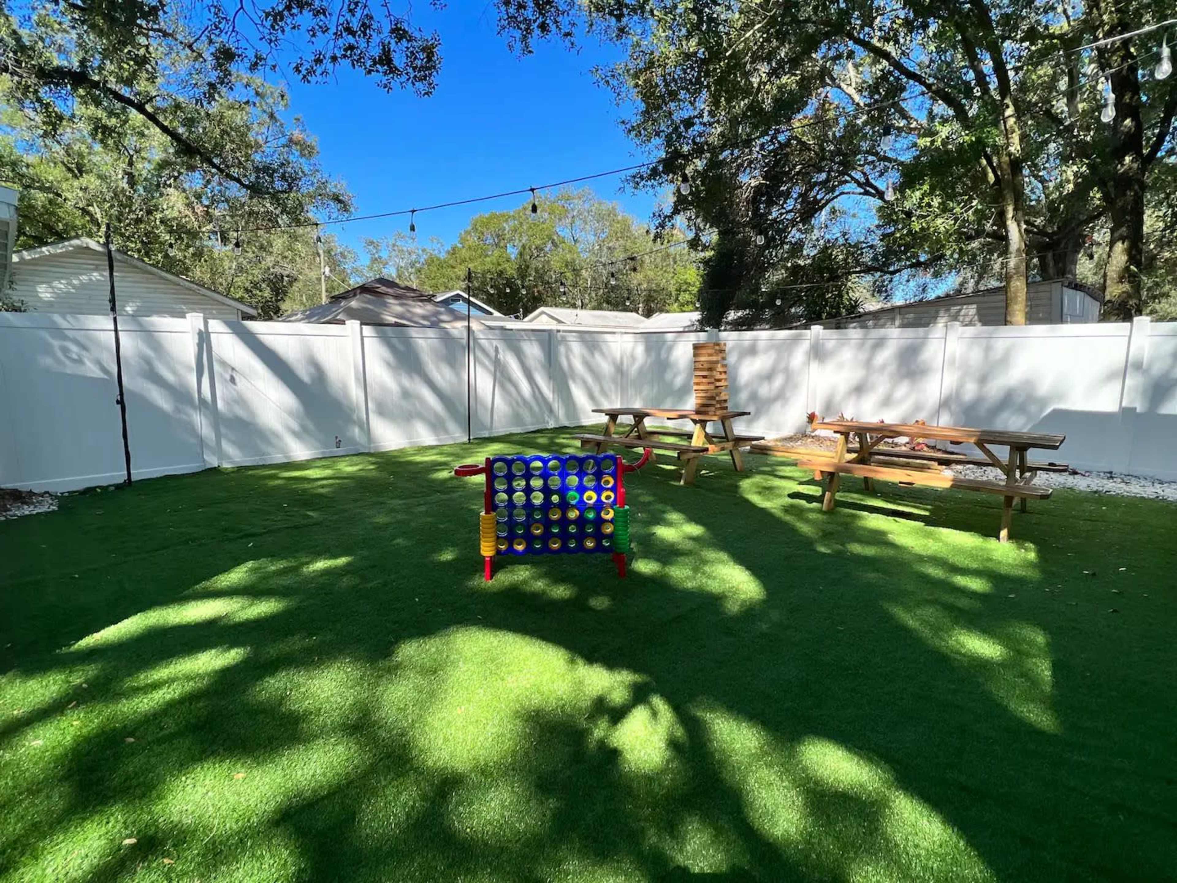 A green artificial lawn area with a large Connect Four game and several wooden picnic tables surrounded by a white fence.