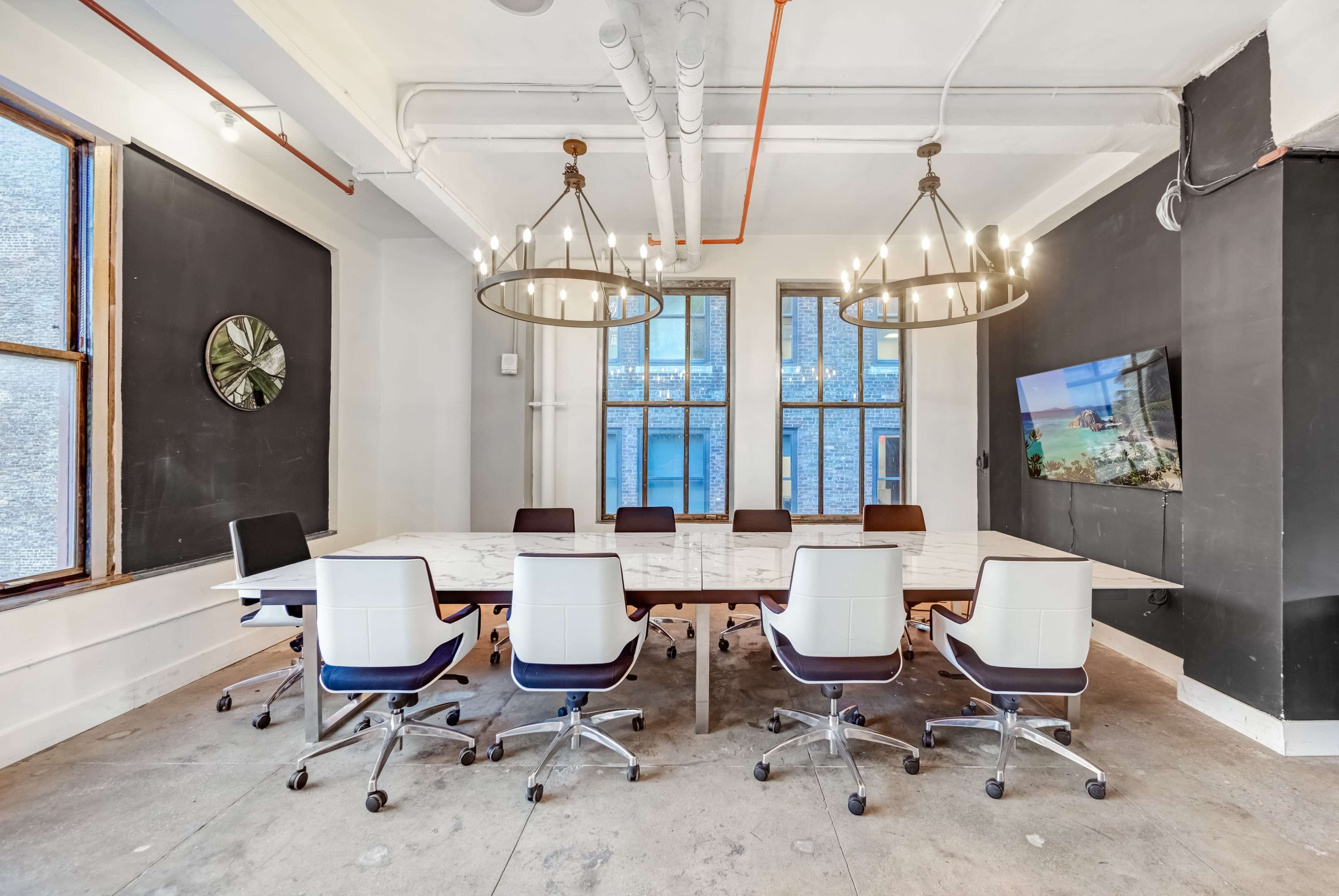 A modern conference room features a large marble table surrounded by white and black chairs, with two chandeliers overhead and a wall-mounted screen.