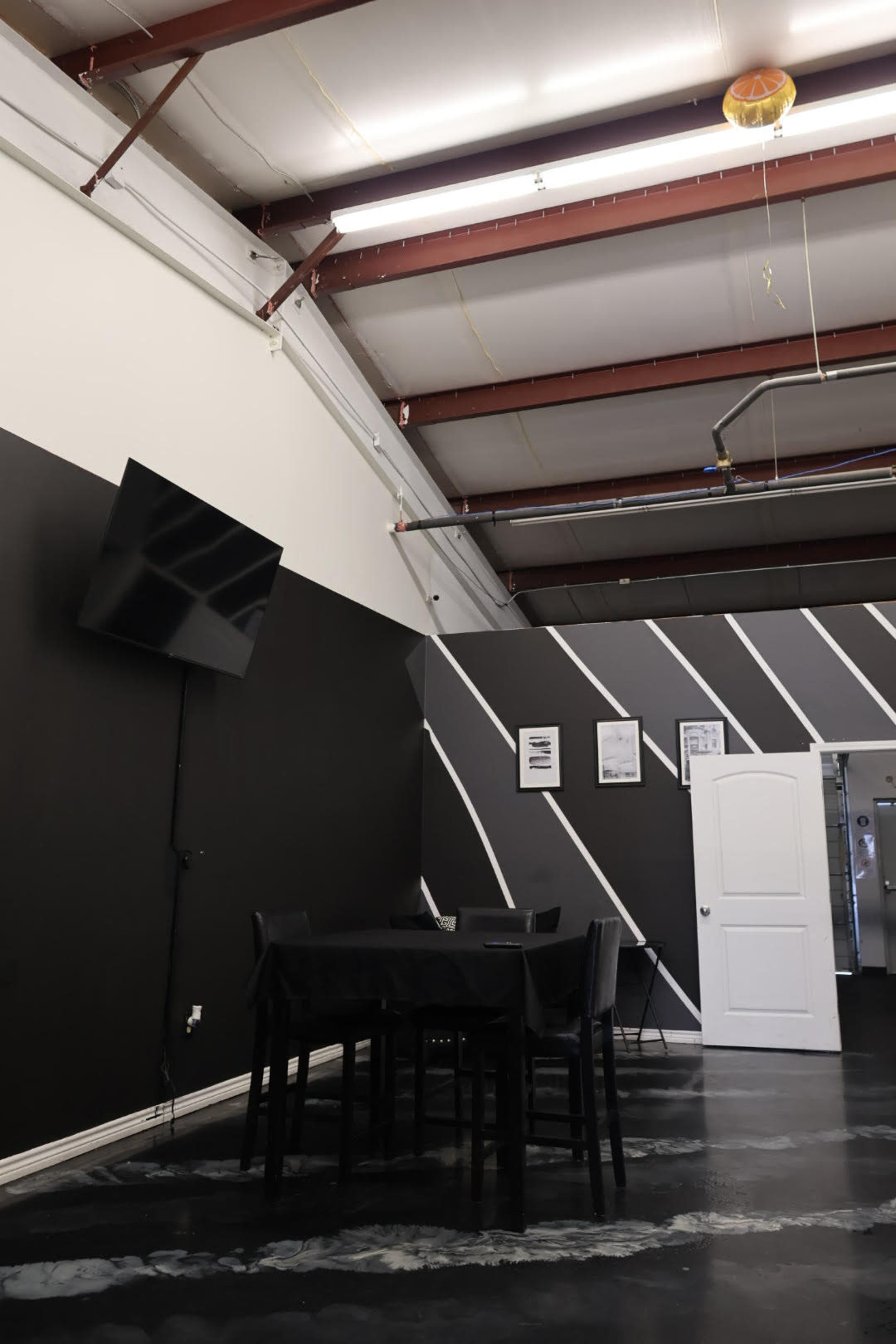 The image shows a spacious interior with black walls, a television mounted on one wall, a table with chairs, and an open door leading to another area.