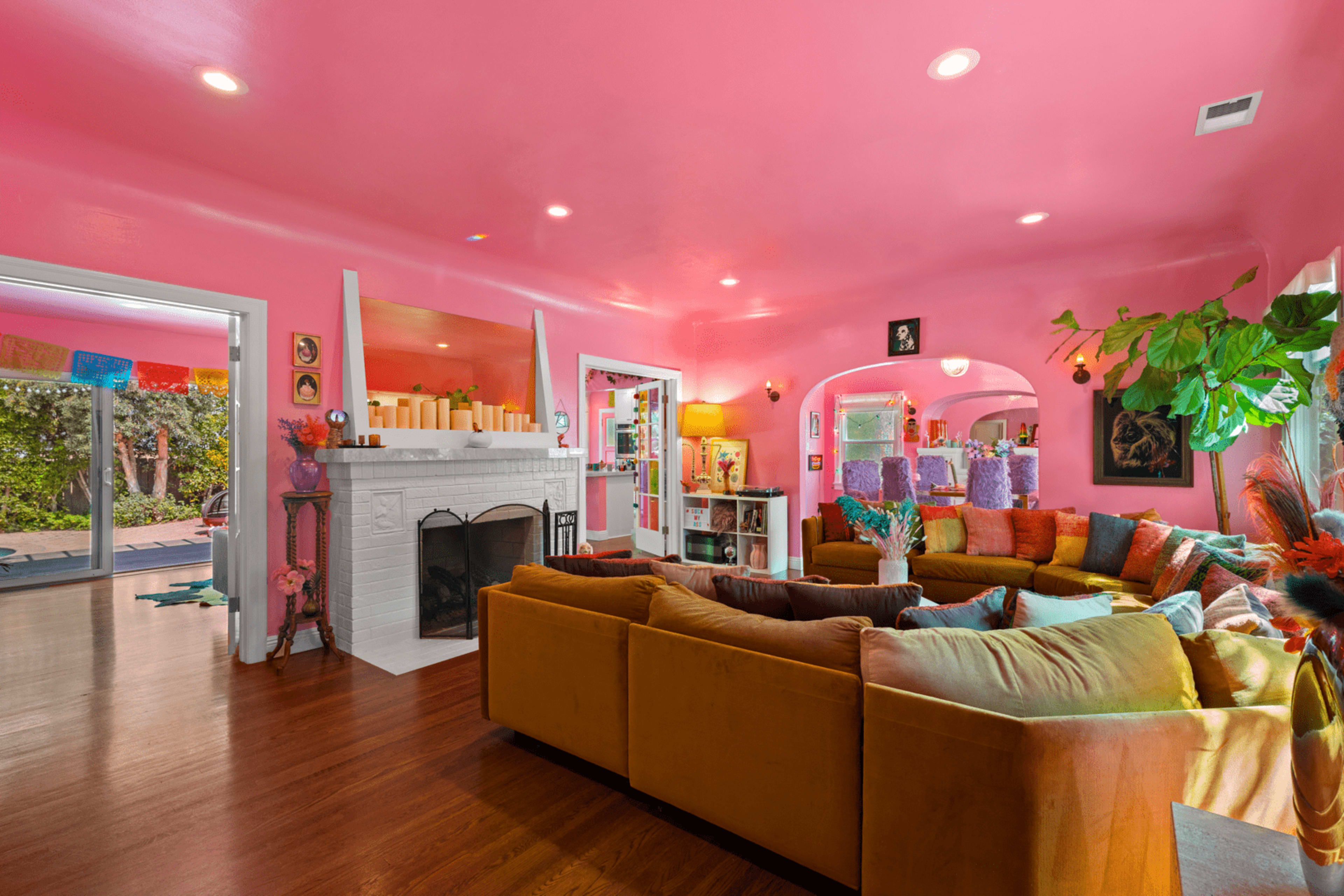 The room features a bright pink wall with a large, comfortable sectional sofa surrounded by various decorative elements and a fireplace.