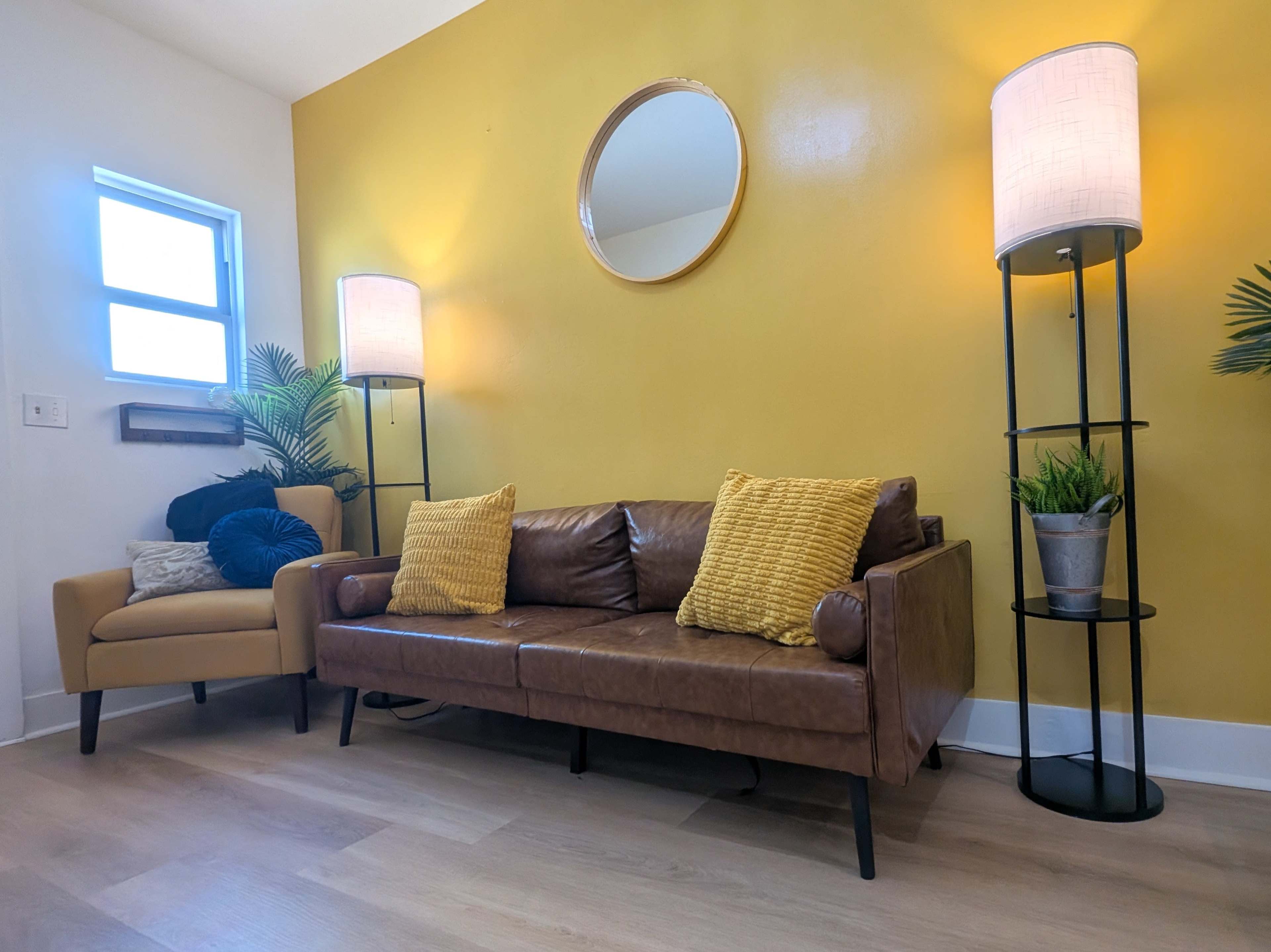 The image shows a cozy living room with a brown leather sofa, a beige armchair, two standing lamps, and a yellow wall decorated with a round mirror.