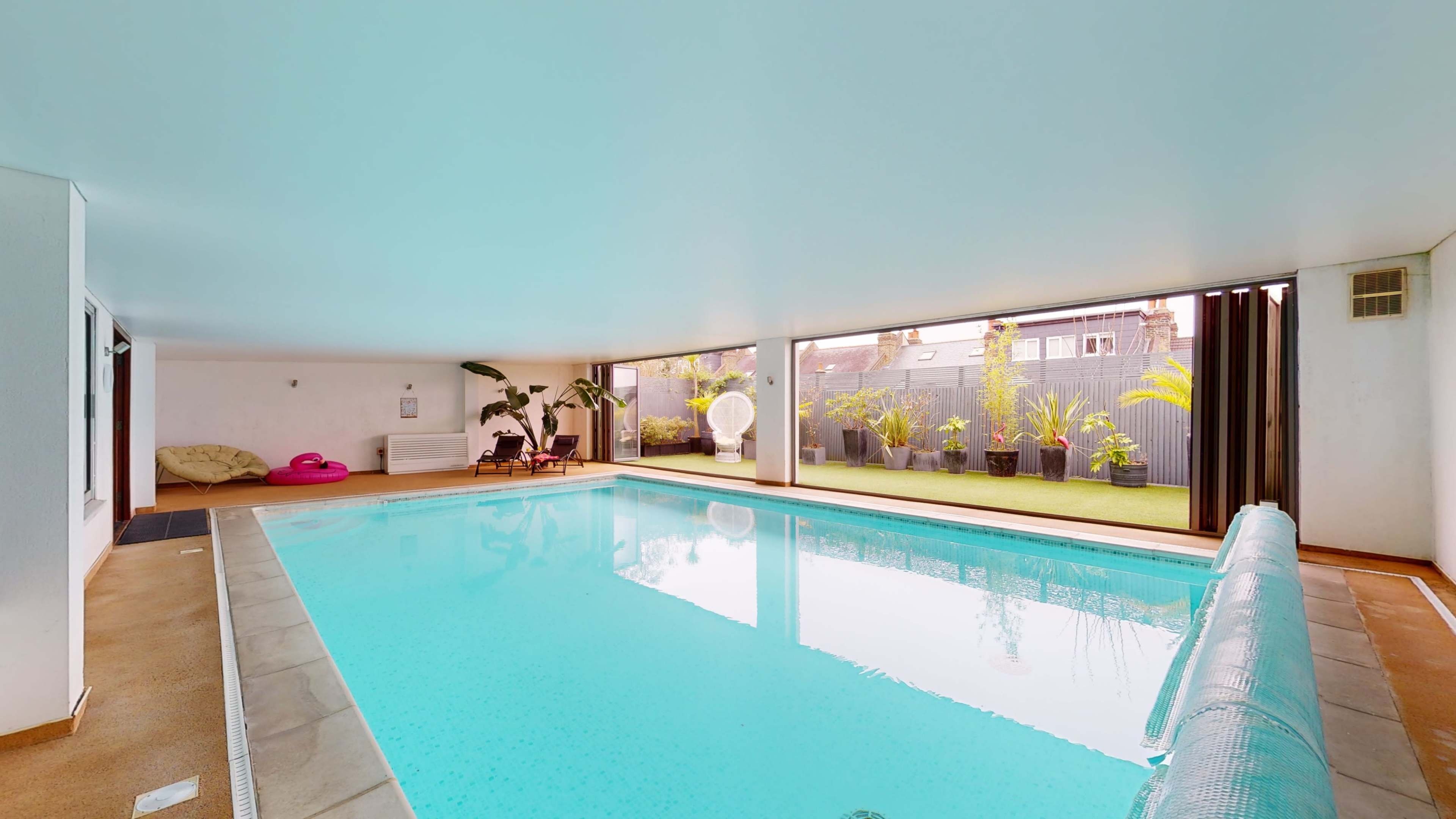 The image shows an indoor swimming pool surrounded by large windows and potted plants.