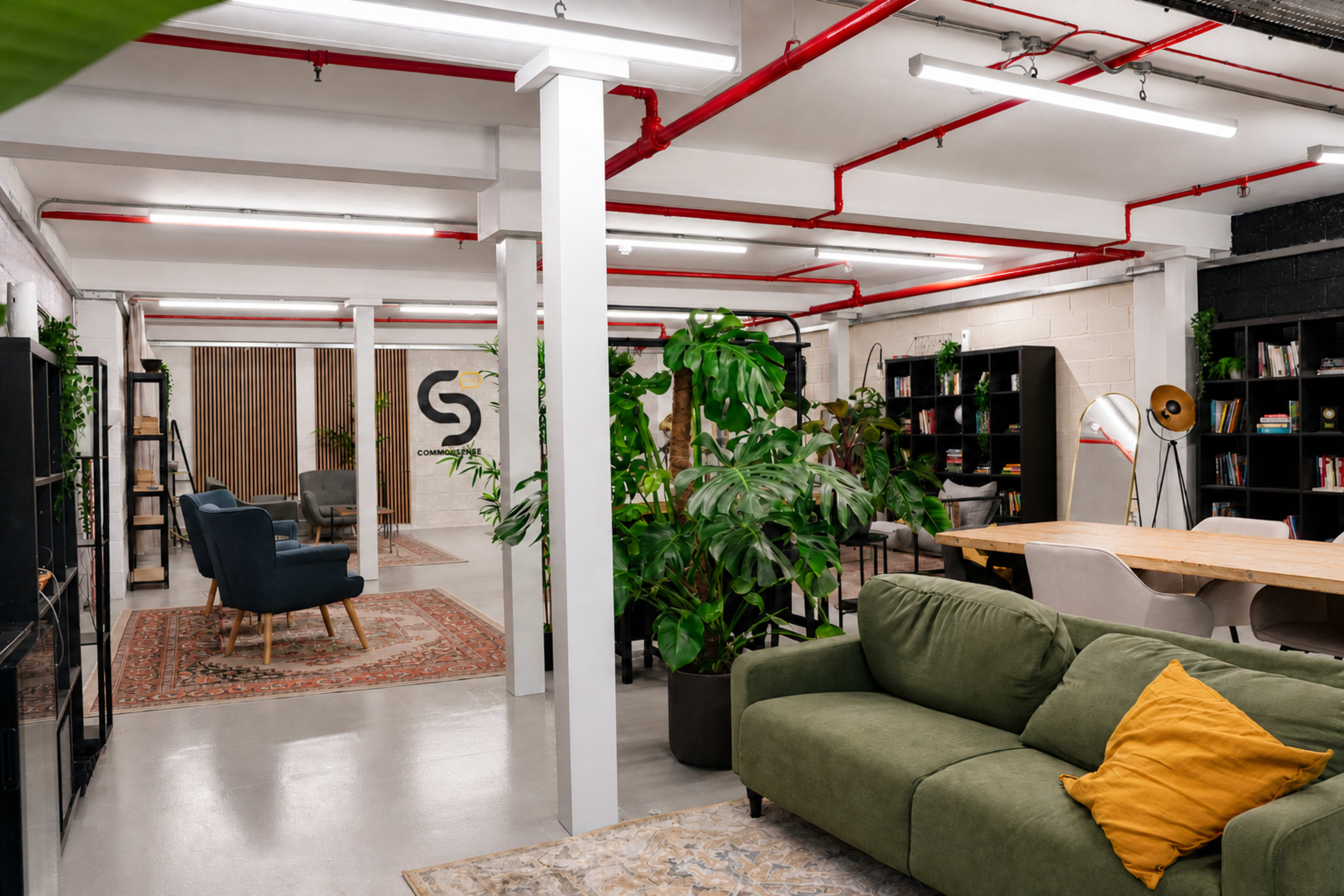 The image shows a modern office space featuring a lounge area with green plants, a large table, comfortable seating, and shelves filled with books.