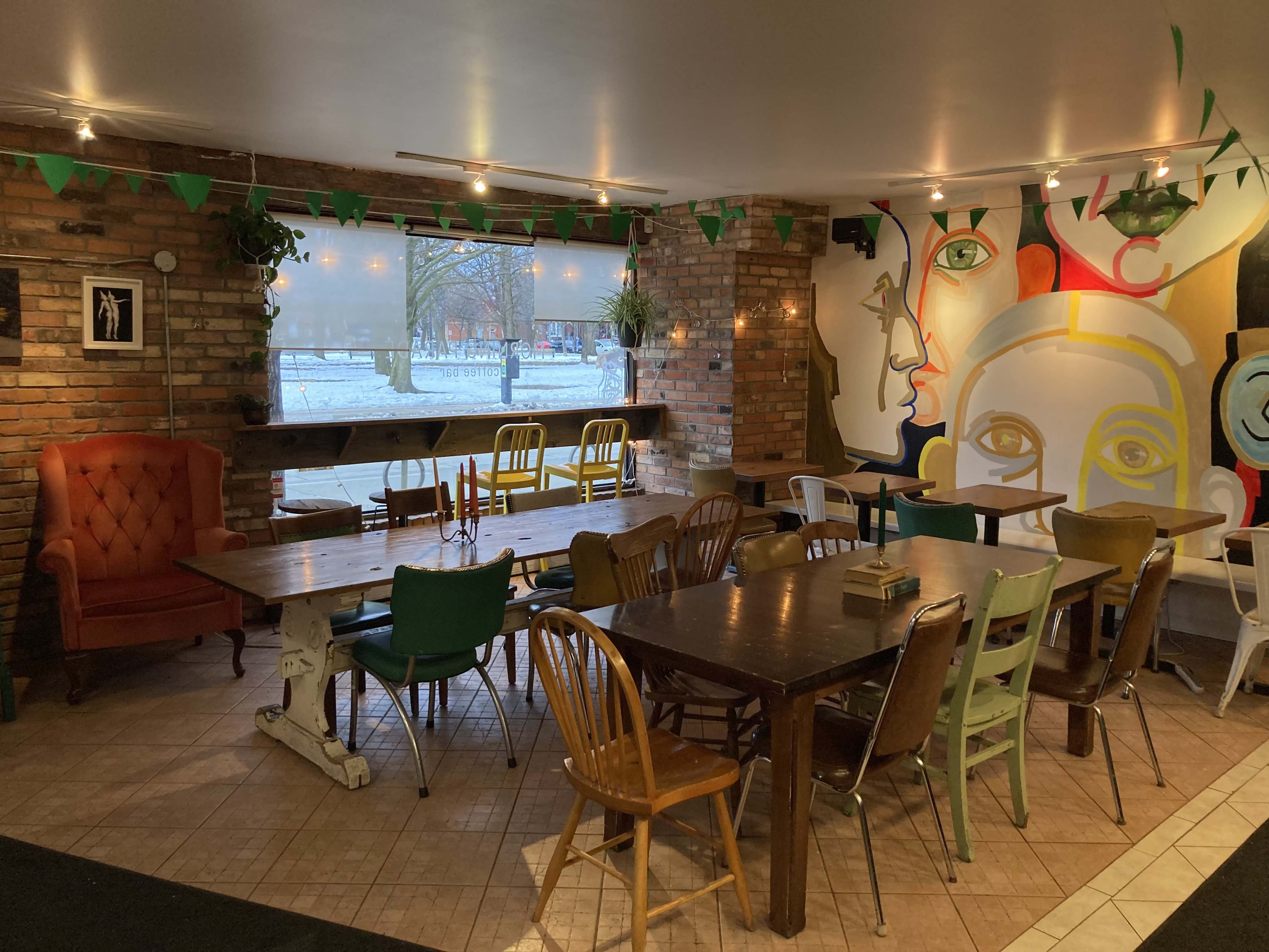 The image shows an interior of a cafe with a mix of wooden and metal tables and chairs, exposed brick walls, and a colorful mural featuring abstract faces.