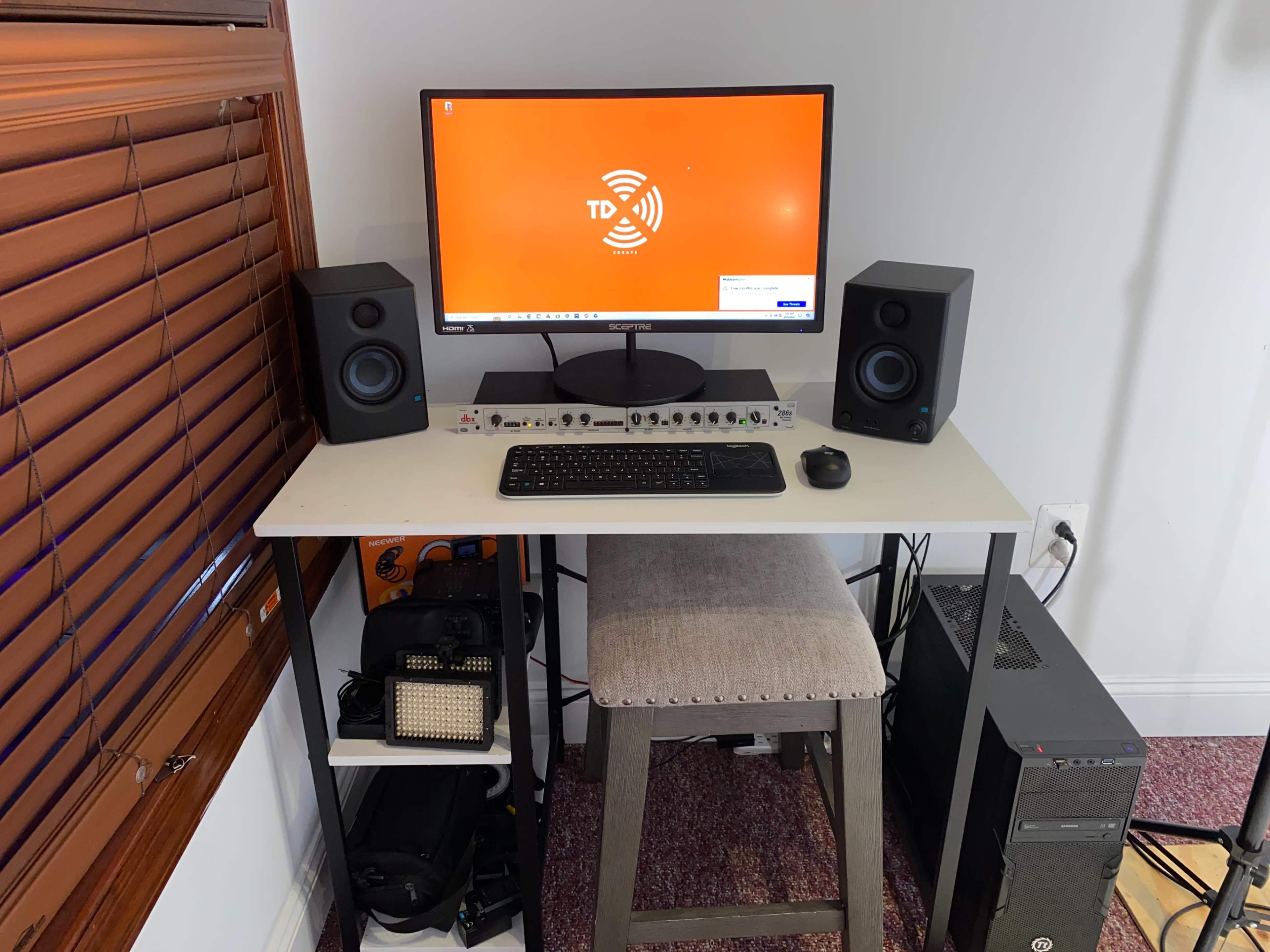 The image shows a computer desk setup featuring a monitor, speakers, a keyboard, a mouse, and various tech equipment, all positioned next to a window with wooden blinds.