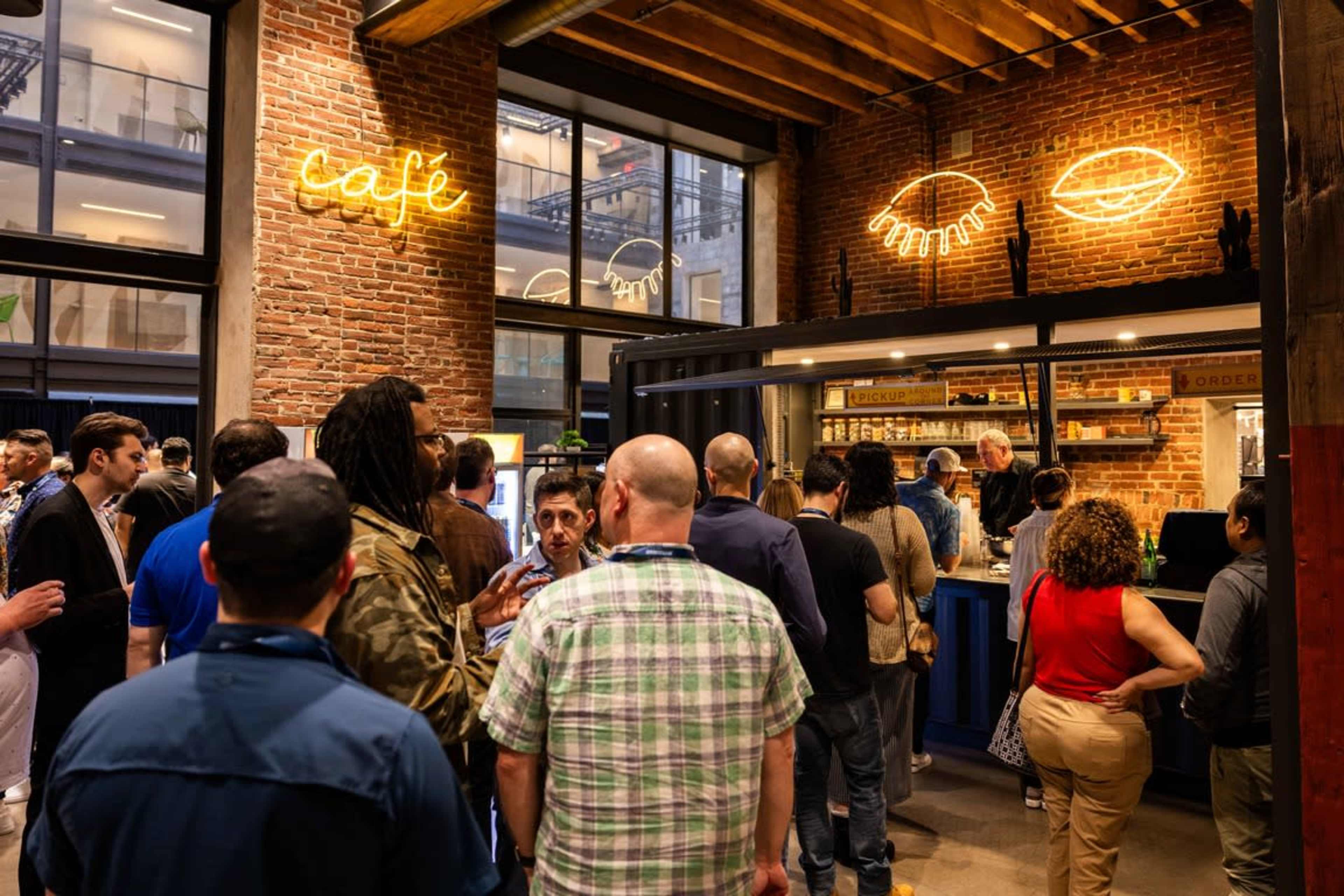 A crowd of people gathers in a cafe with exposed brick walls and neon signage.