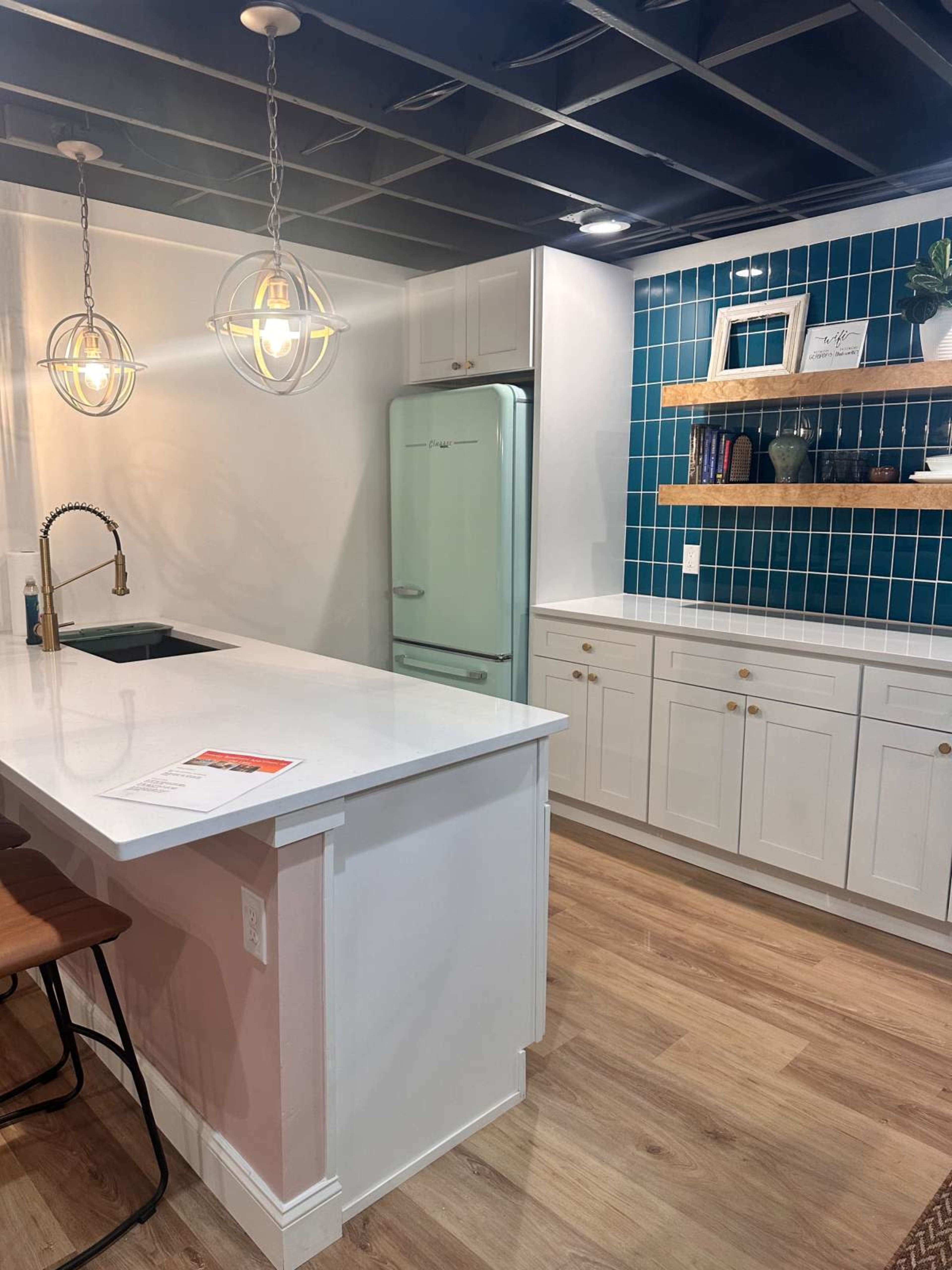 The image shows a modern kitchen with a white countertop, teal tiled wall, open shelves, and vintage-style light fixtures.