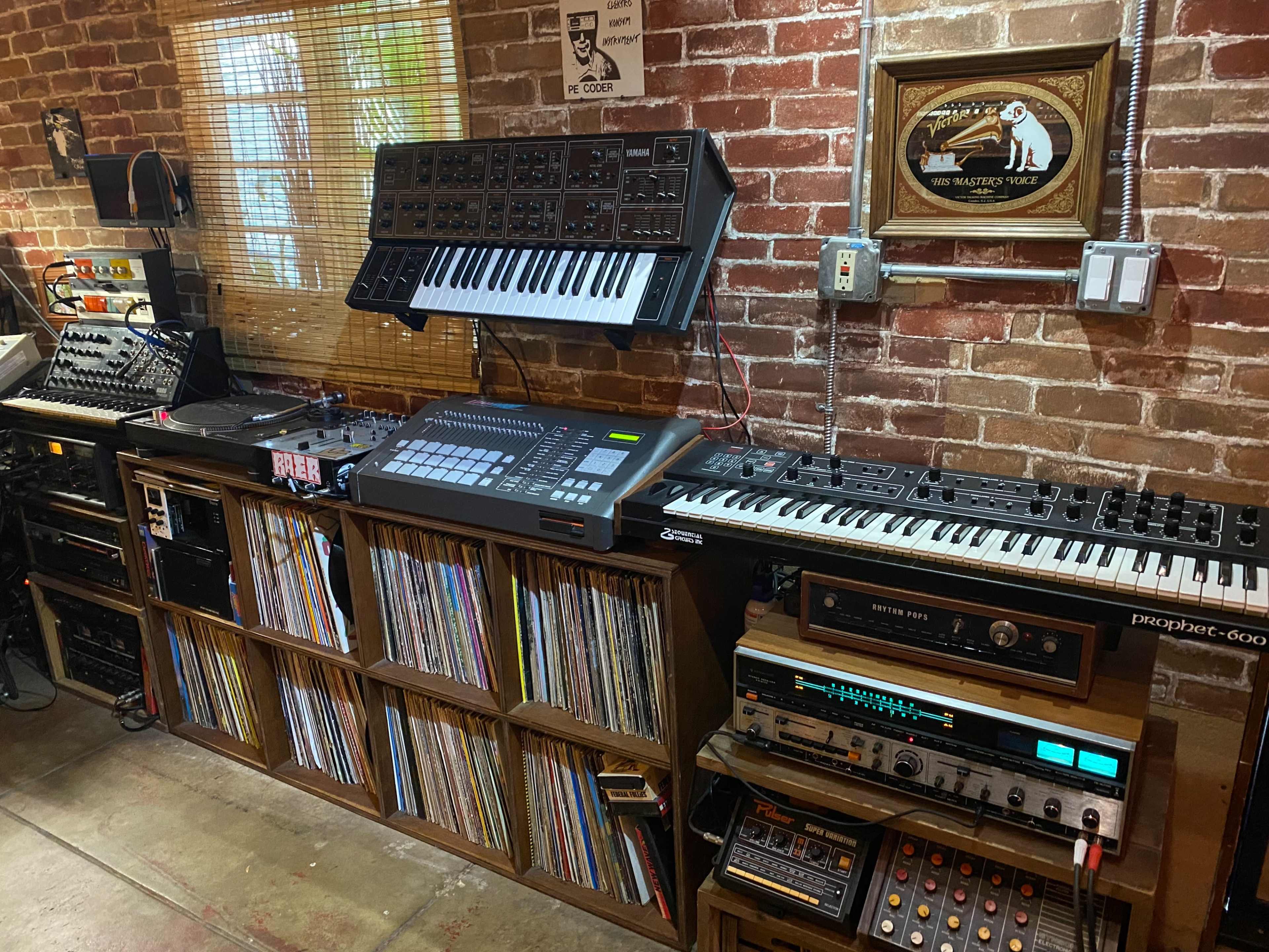 A music production setup featuring synthesizers, a digital audio workstation, vinyl records, and an audio mixer against a brick wall.