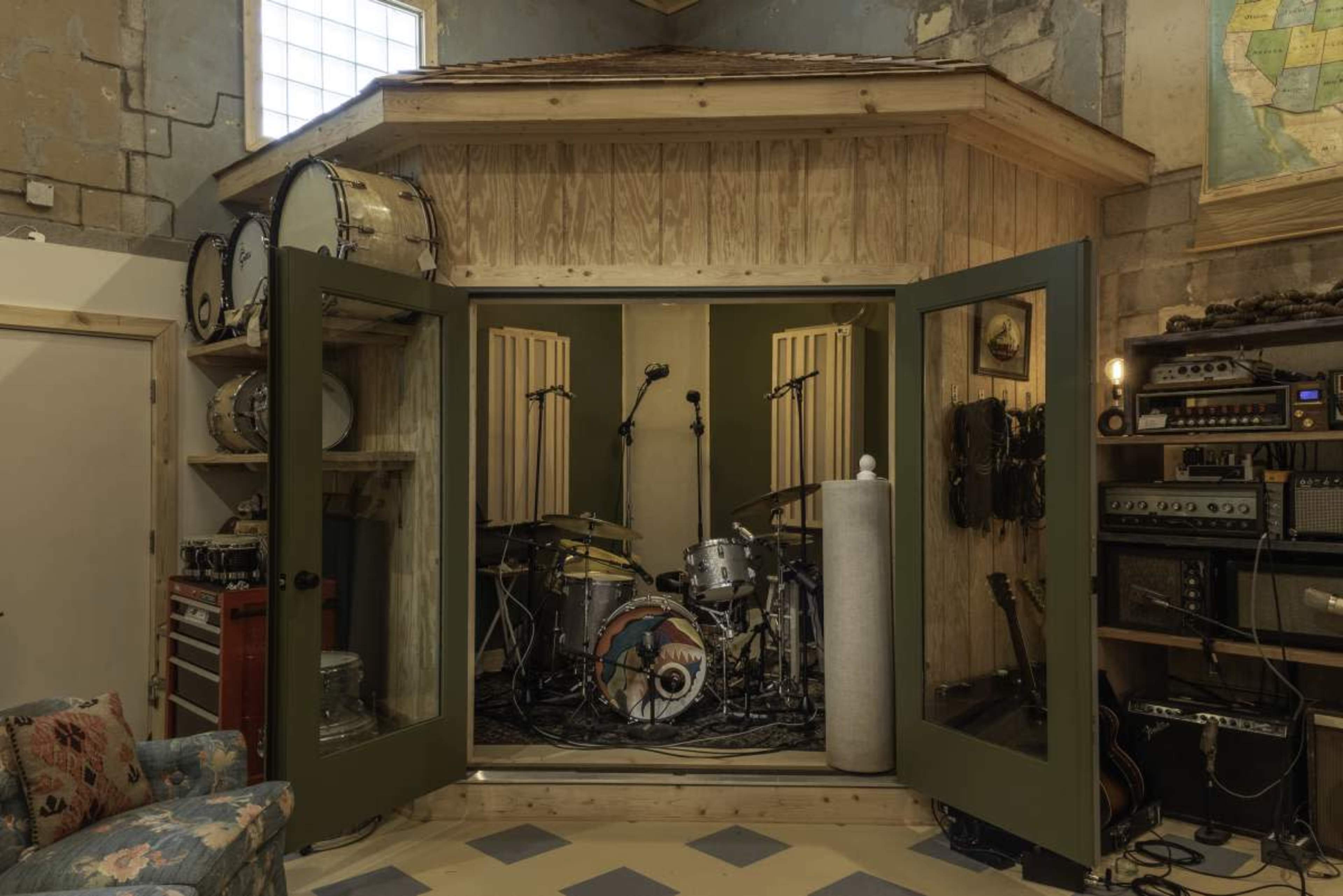 The image shows a wooden sound booth with open doors, revealing a drum set and audio equipment inside.