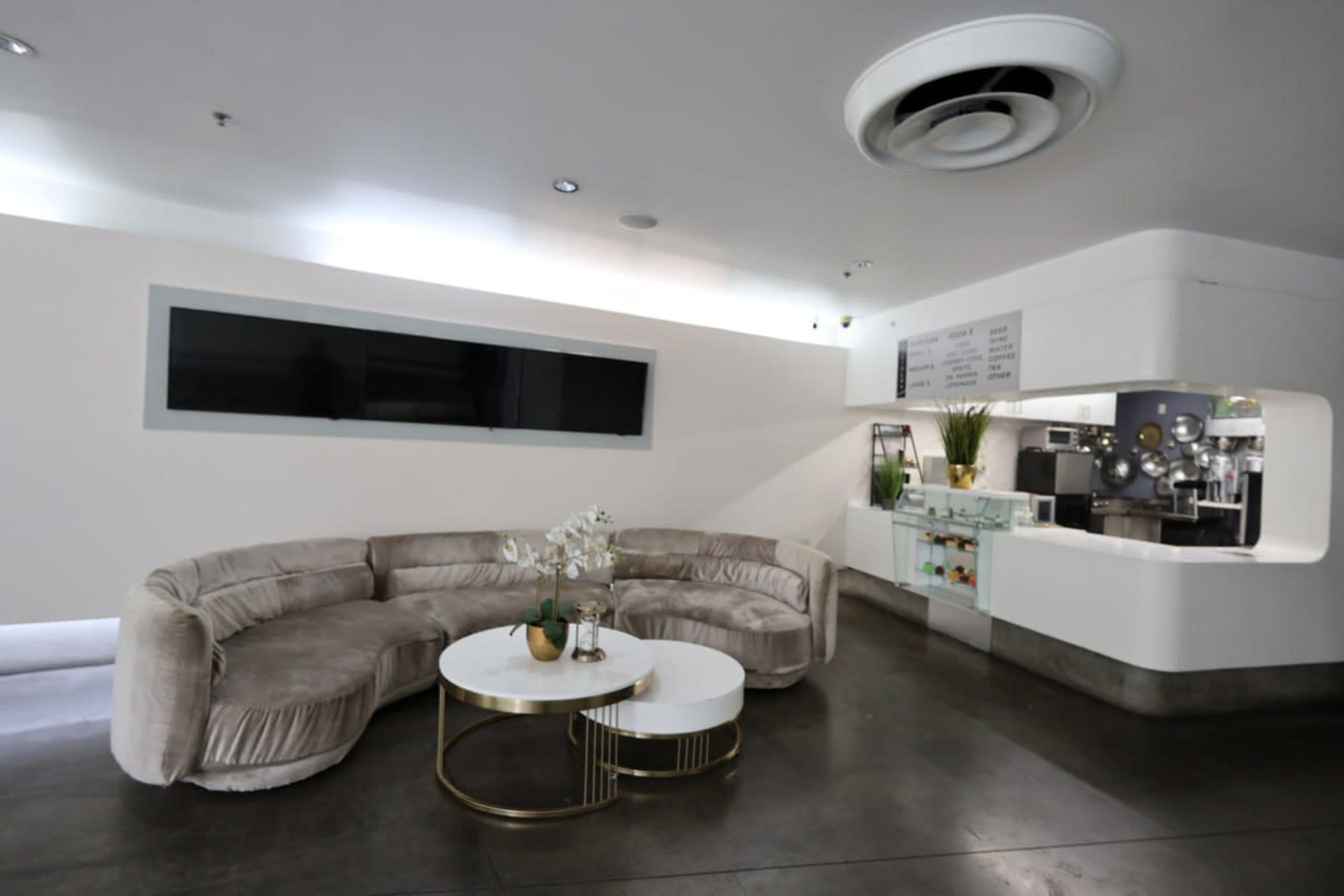 The image shows a modern, minimalist lounge area with a curved sofa, a small round coffee table, and a service counter in a bright, spacious room.