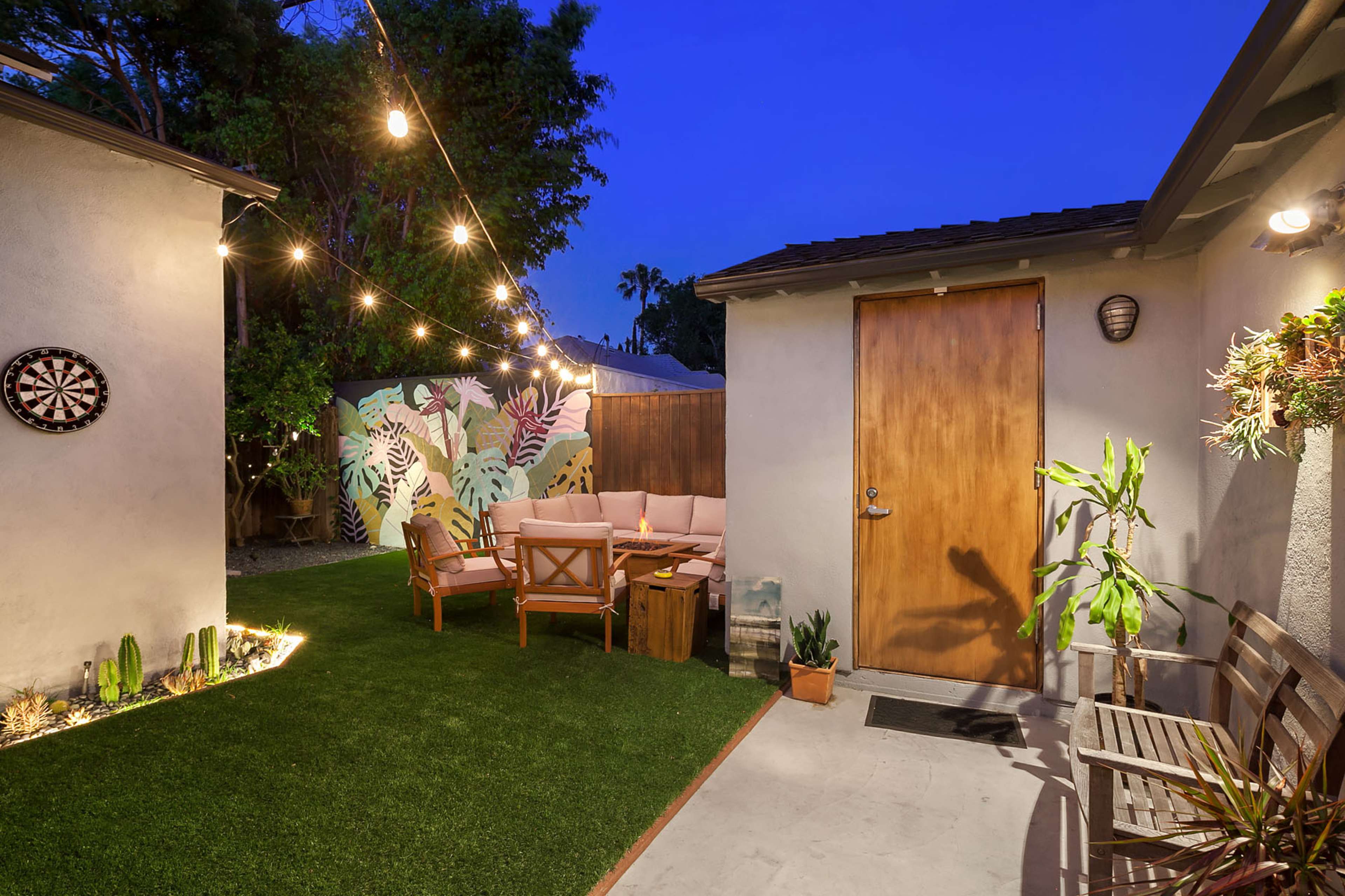 The scene features a backyard patio with a cozy seating arrangement, decorative plants, a dartboard, and string lights illuminating the area at dusk.