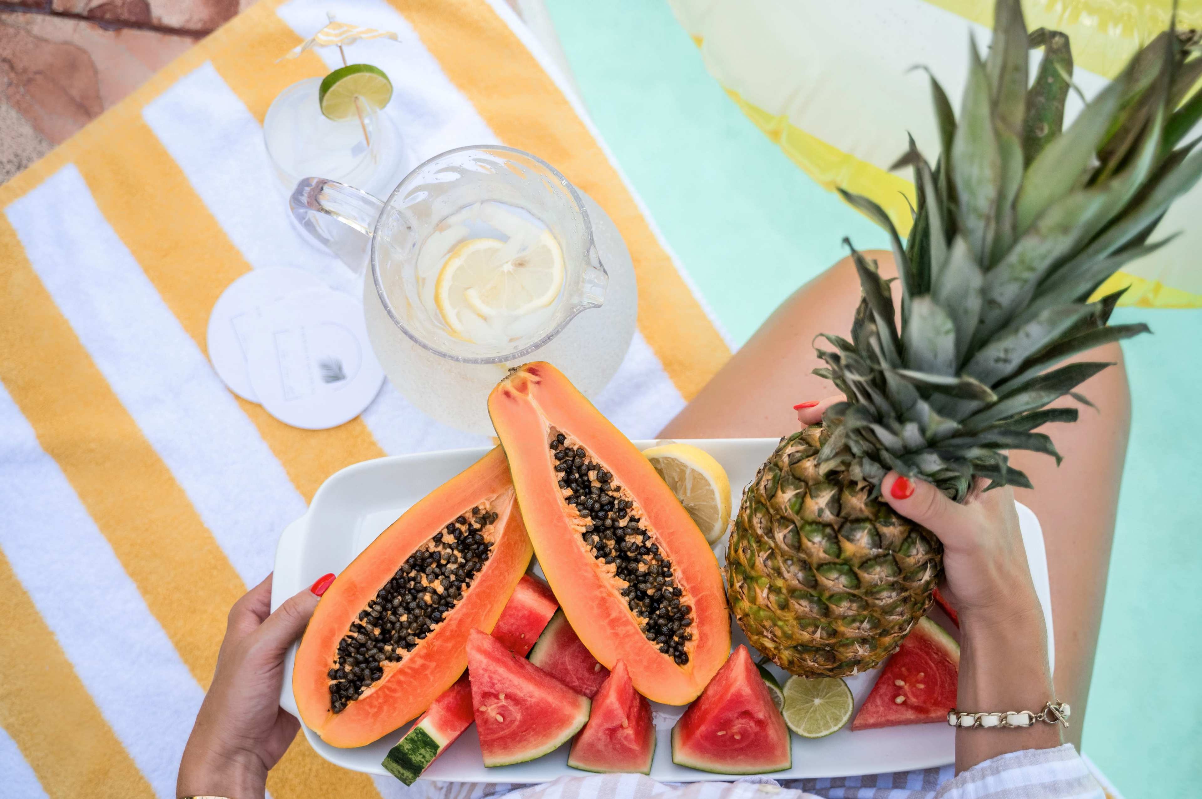 A person holds a tray with sliced papaya, watermelon, and a pineapple beside a pitcher of lemonade on a towel.