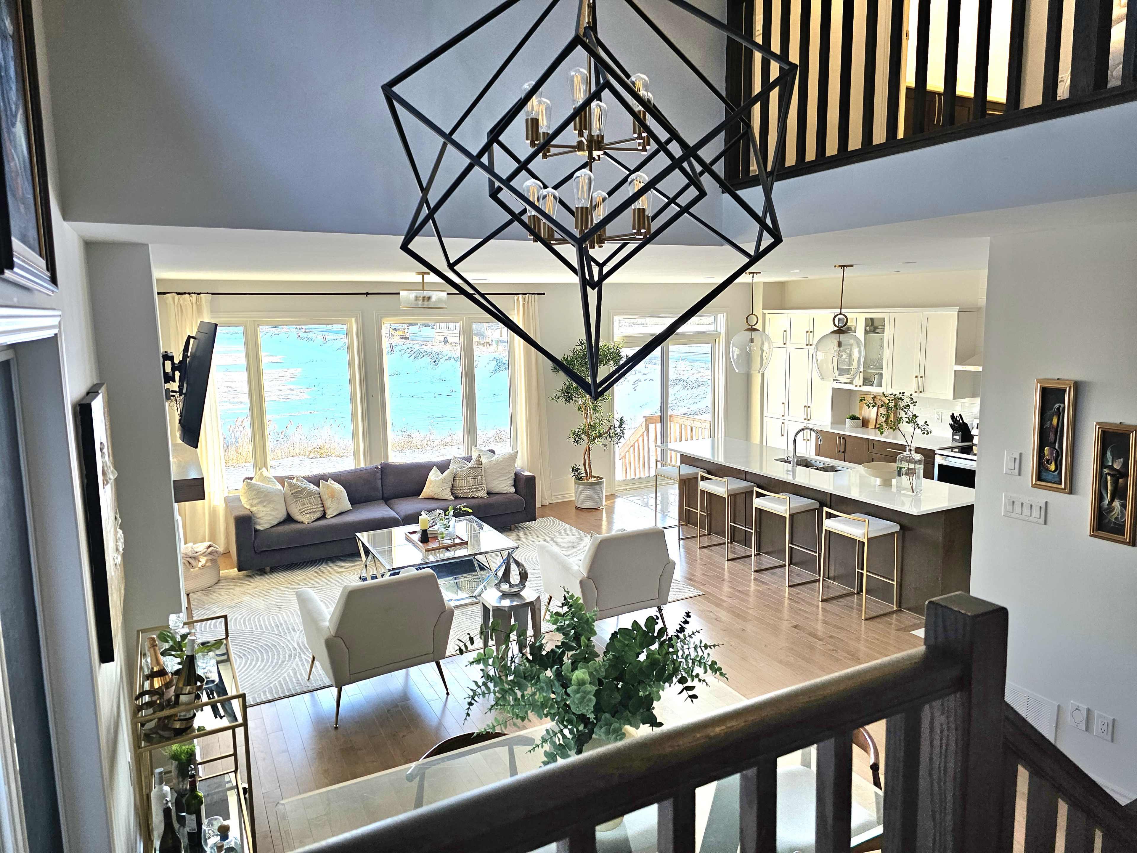 A modern living room with a view of water, featuring a gray sofa, white chairs, a coffee table, and a large chandelier overhead.