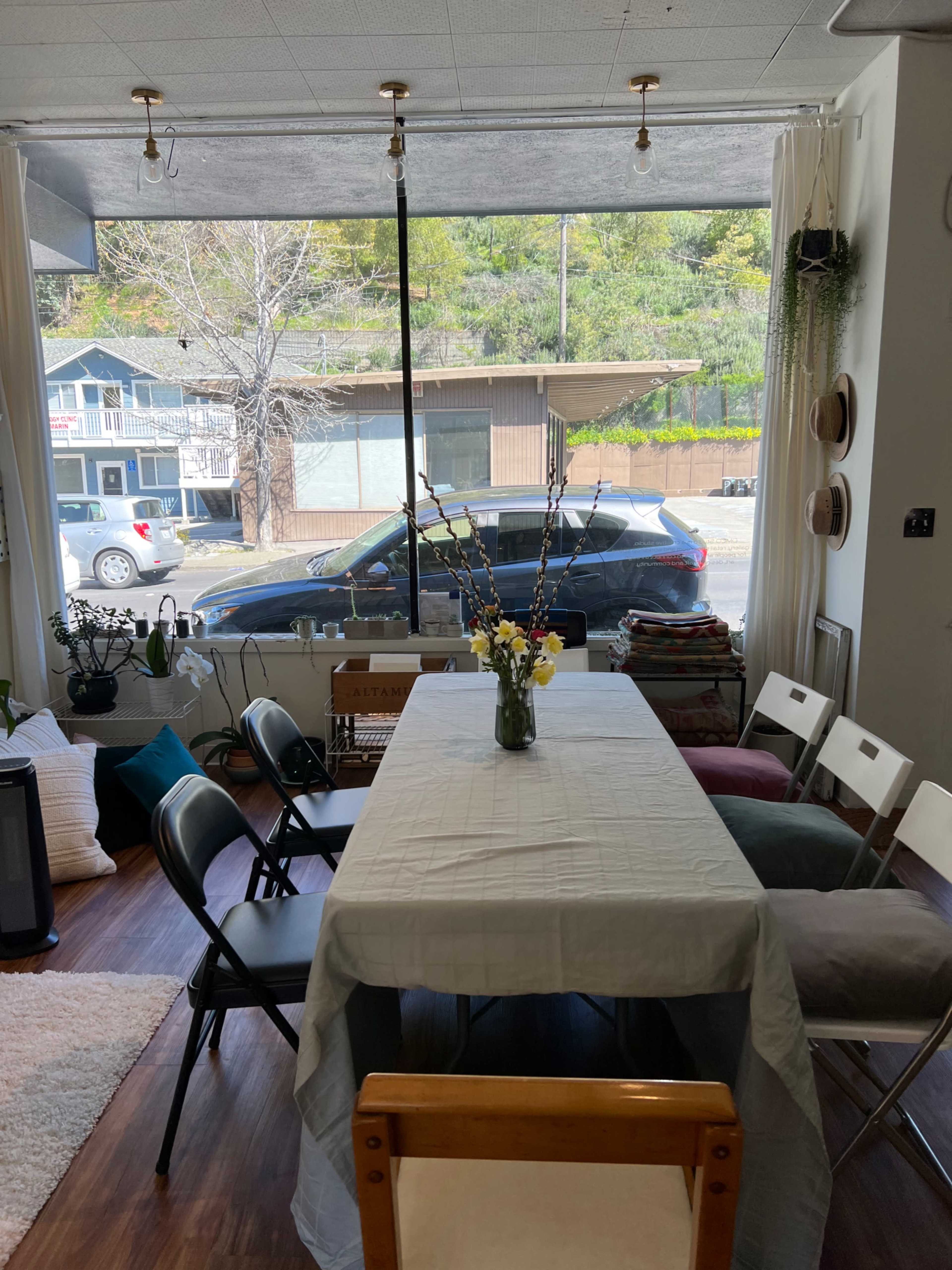 A long table with a tablecloth and a vase of flowers is set up in front of a large window facing a street with parked cars.