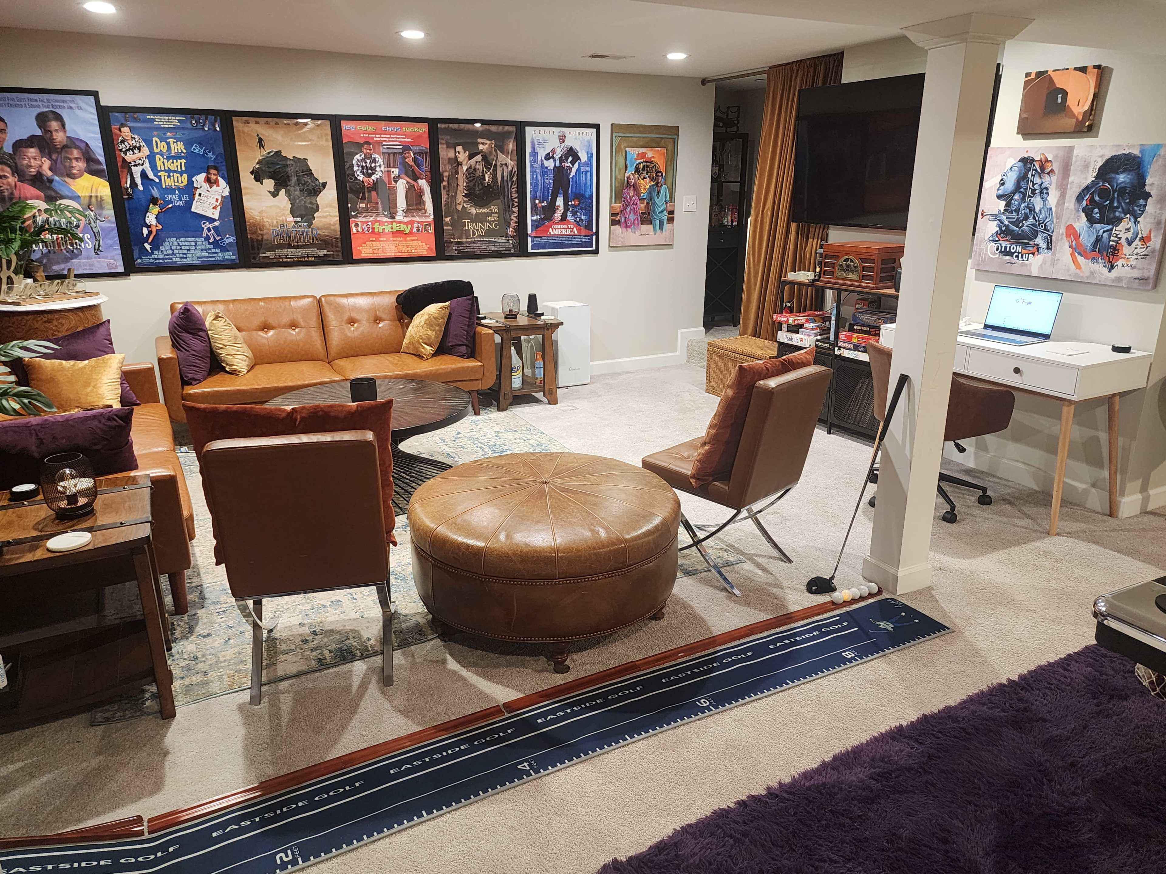The image shows a cozy basement living area featuring a brown leather sofa, a round coffee table, various movie posters on the walls, and a workspace with a computer desk.