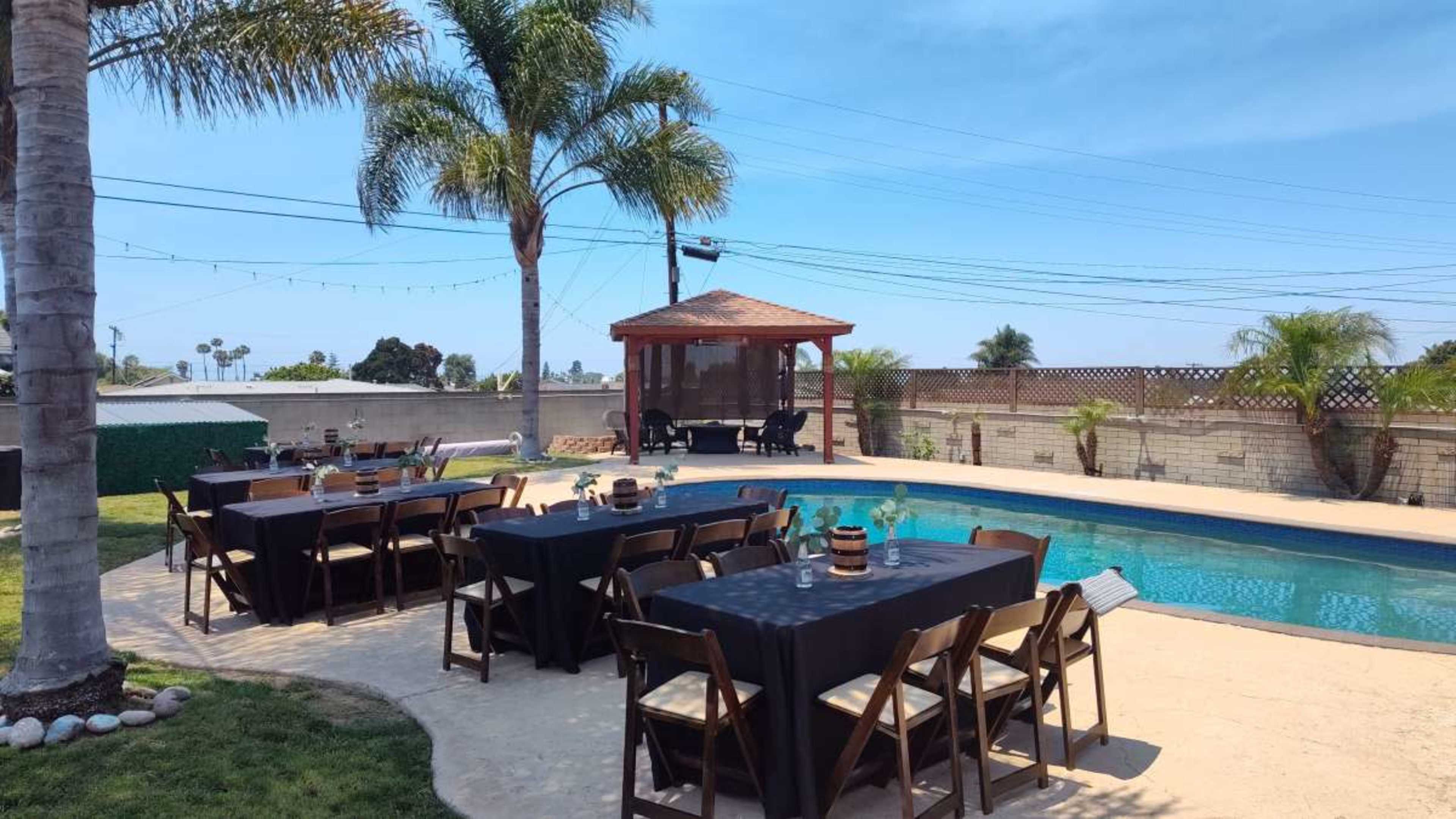 The image shows a poolside area with several black tables and chairs arranged near a sparkling blue pool, surrounded by palm trees and a gazebo.
