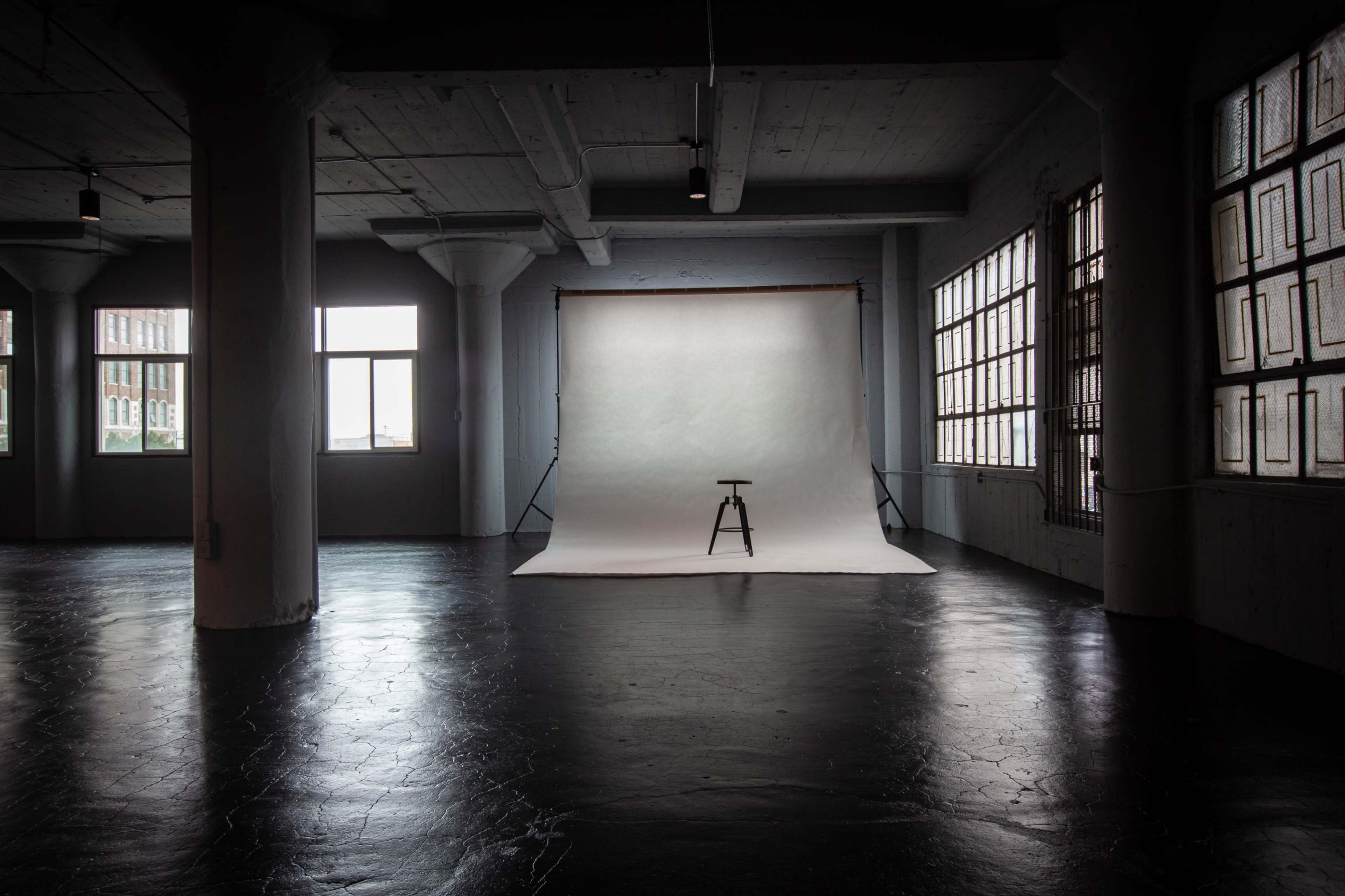 The image depicts an empty photo studio with a white backdrop, a stool in front of it, and large windows allowing natural light to enter.