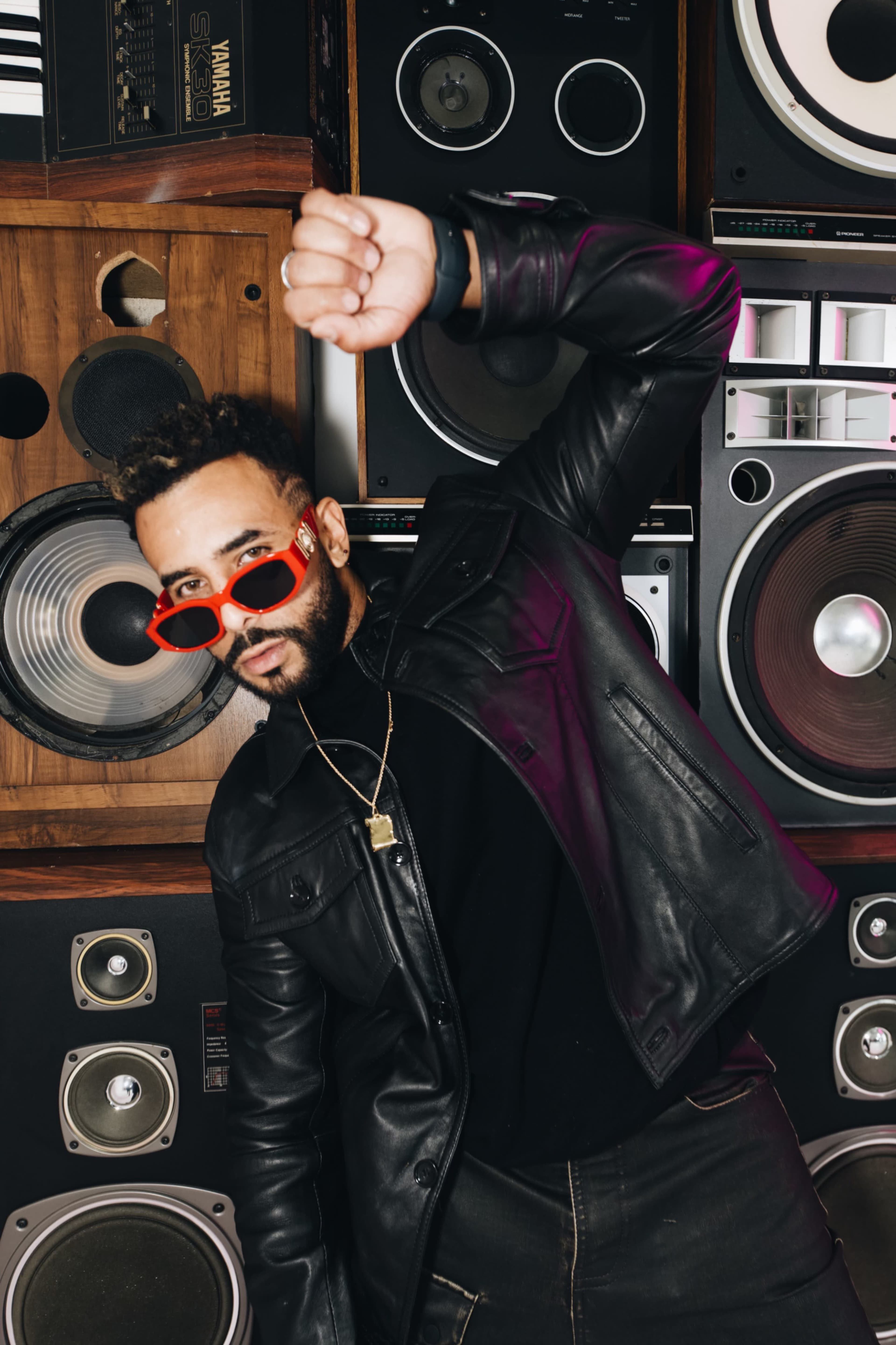 A person in a black leather jacket and red sunglasses poses in front of a wall of vintage speakers.