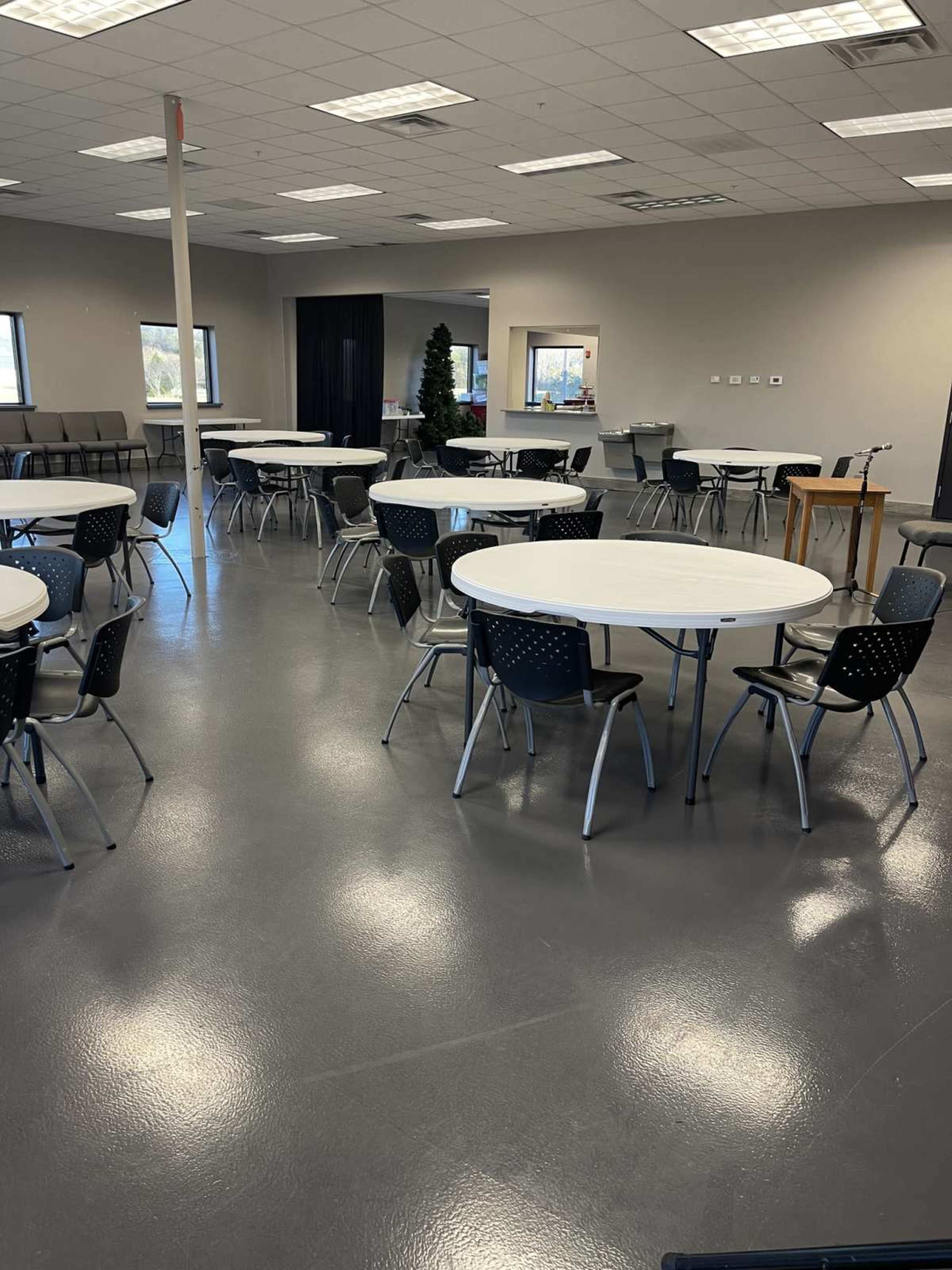 The image shows a large empty room with multiple round tables surrounded by chairs and a few additional seating areas in the background.